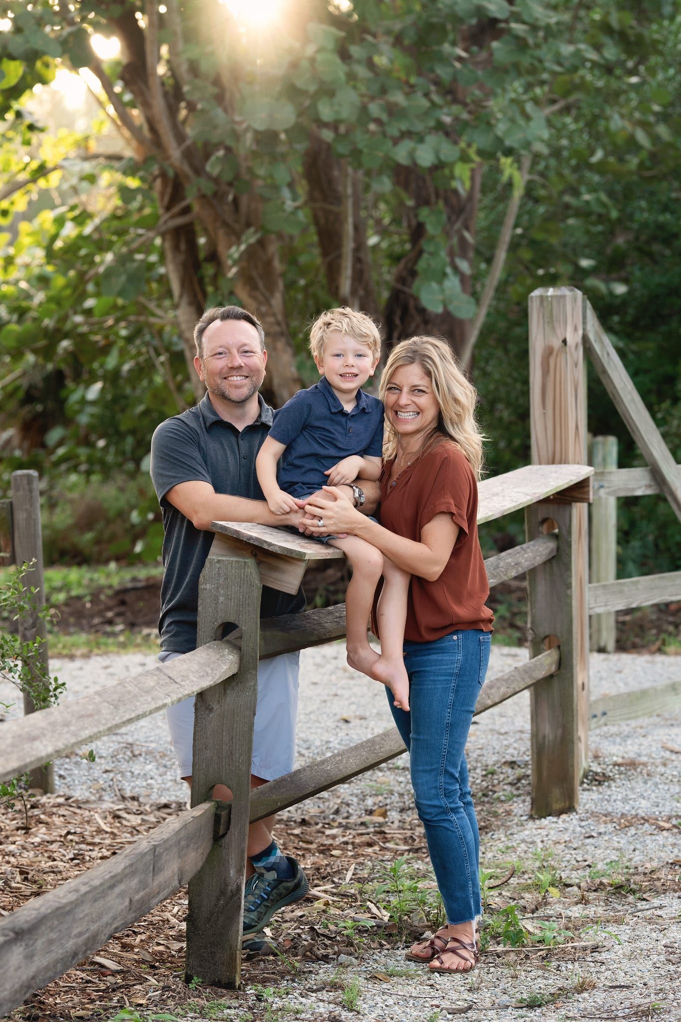 Family of three, smiling, poses on a wooden fence in a natural setting.