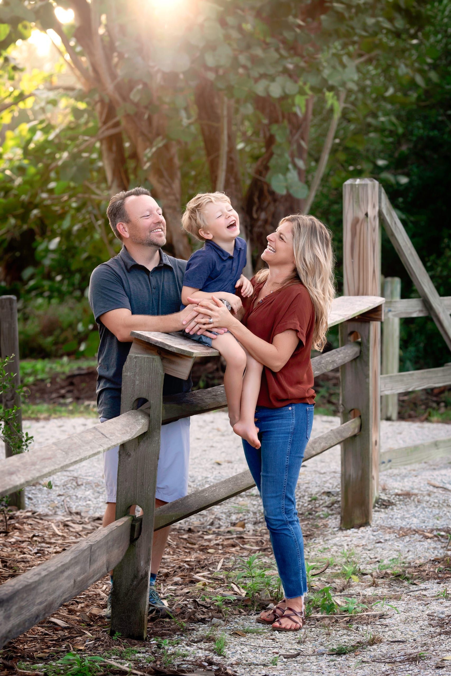 Family laughs together by a wooden fence; sunny outdoor setting.