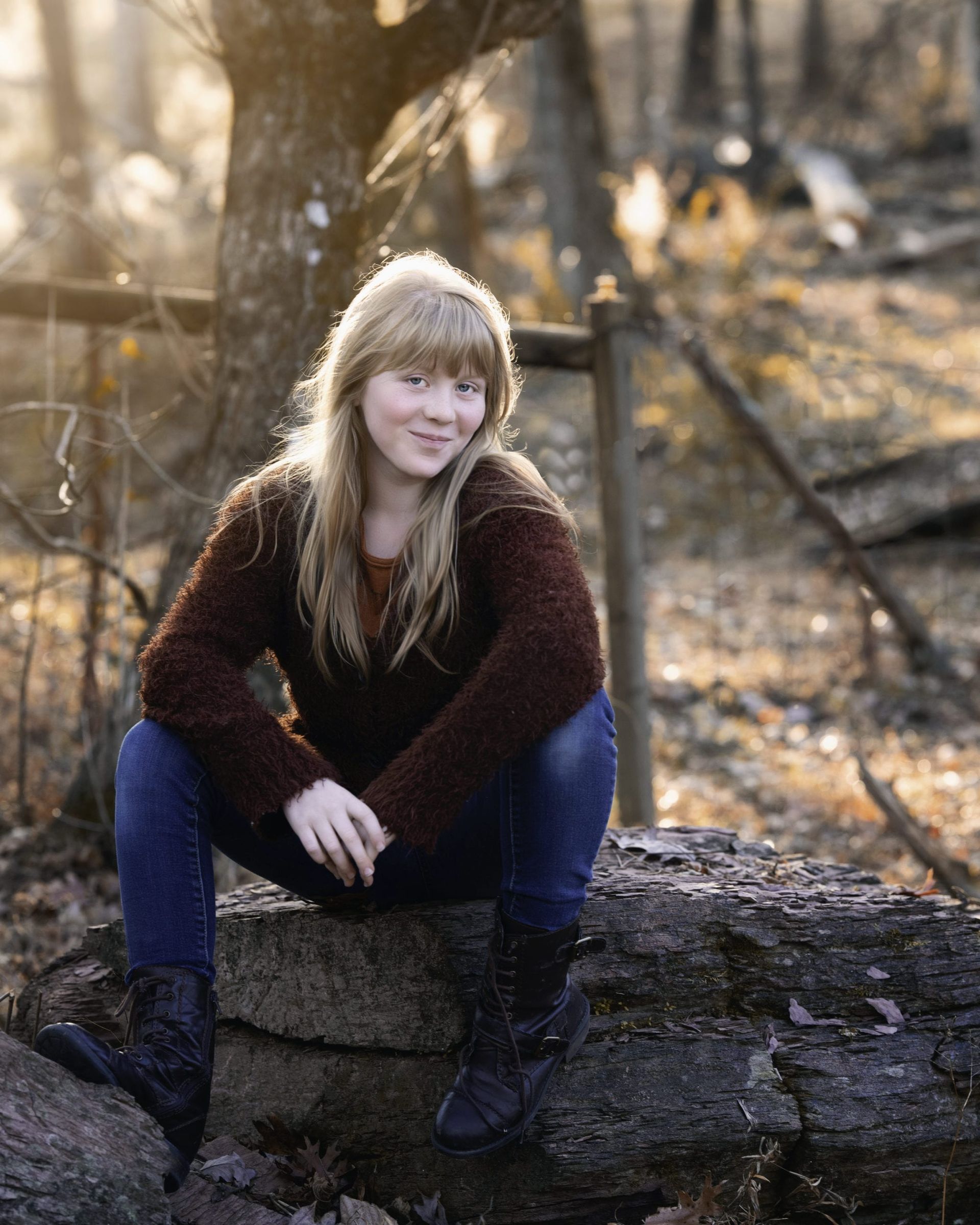 Woman with blonde hair, in a brown sweater and jeans, sits on a log in a sunlit forest, smiling.