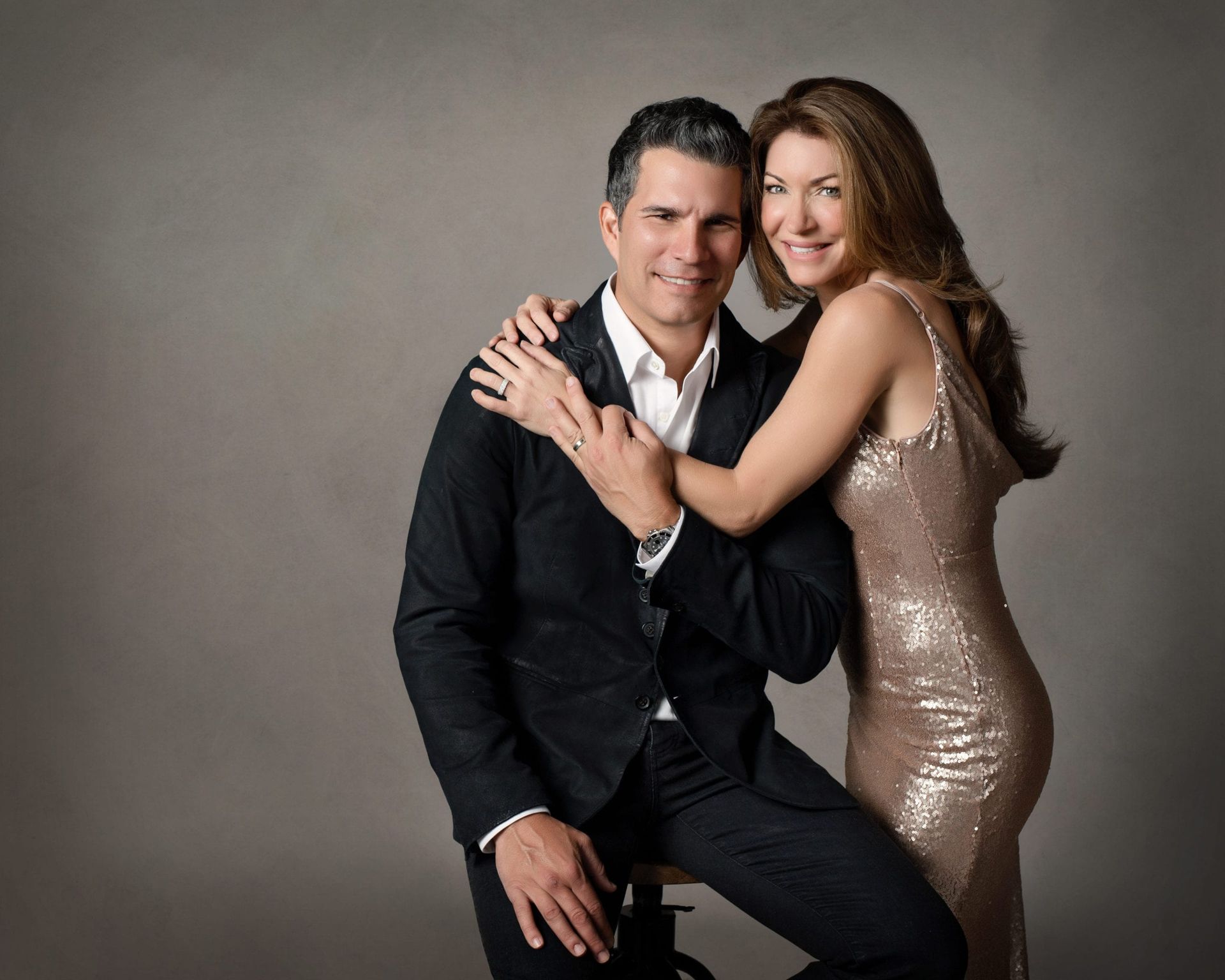 A man and woman pose for a photo. The woman embraces the man. Neutral background.
