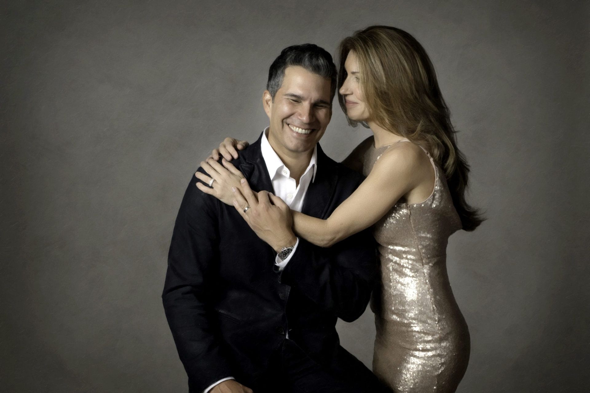 Man in suit smiles, embracing woman in gold sequin dress; neutral background.