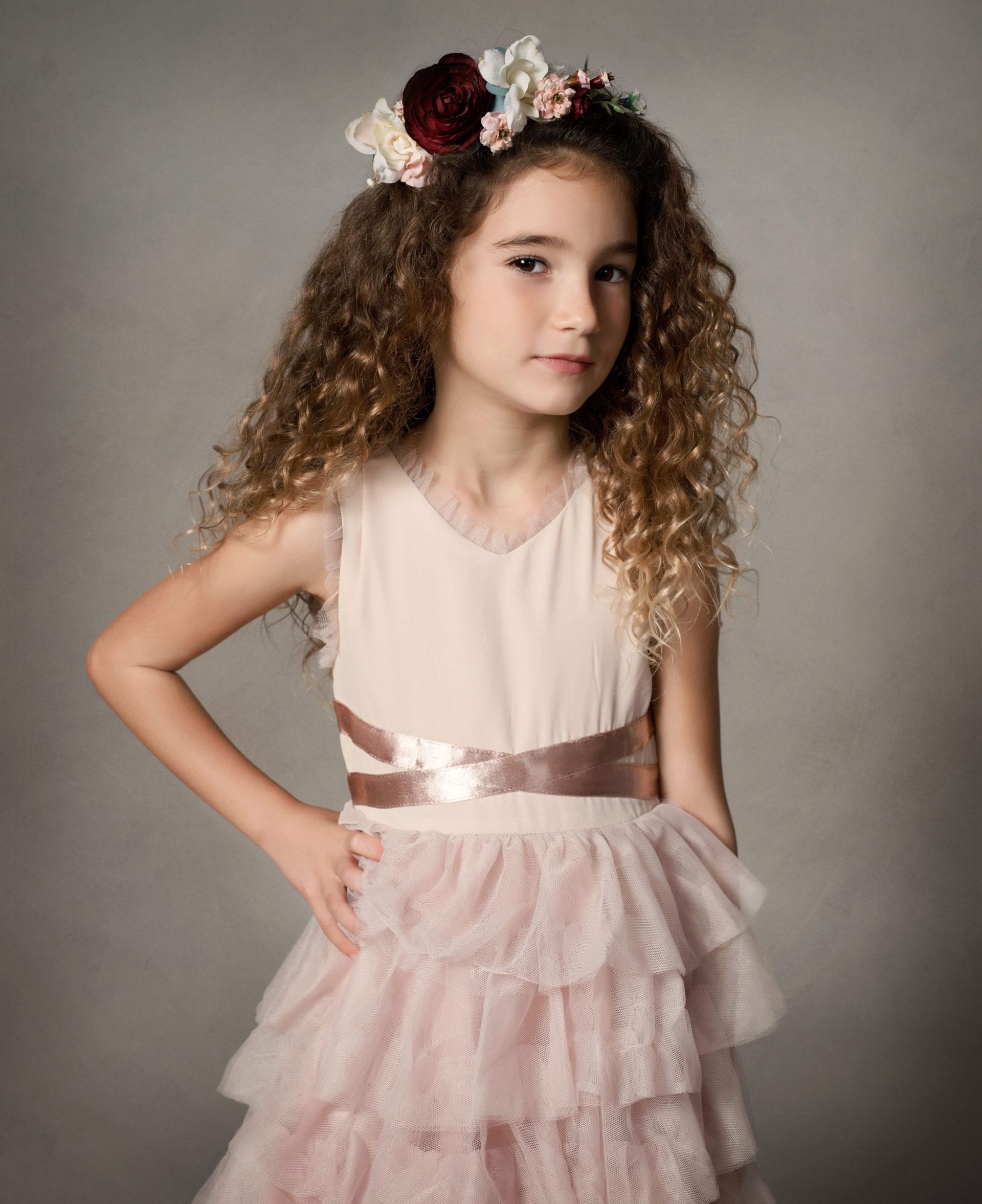 Girl with curly hair wearing a pink dress, flower crown, and posing in front of a gray background.