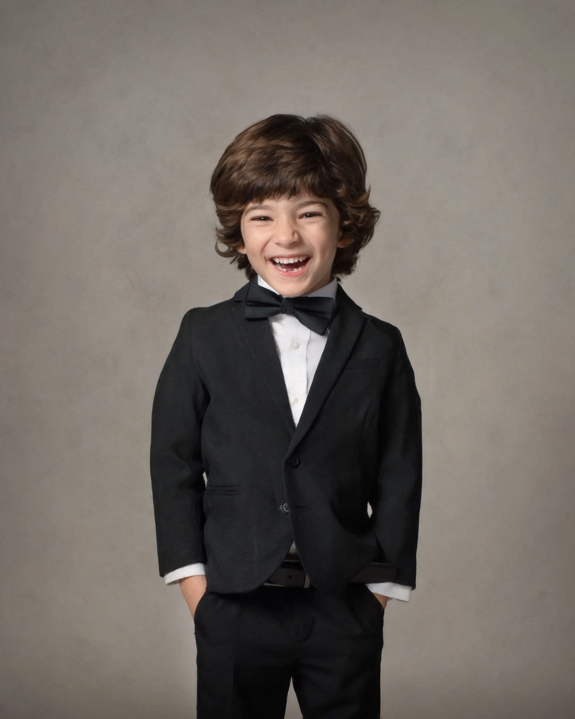Young boy with curly brown hair smiles, wearing a black tuxedo with bow tie, hands in pockets.
