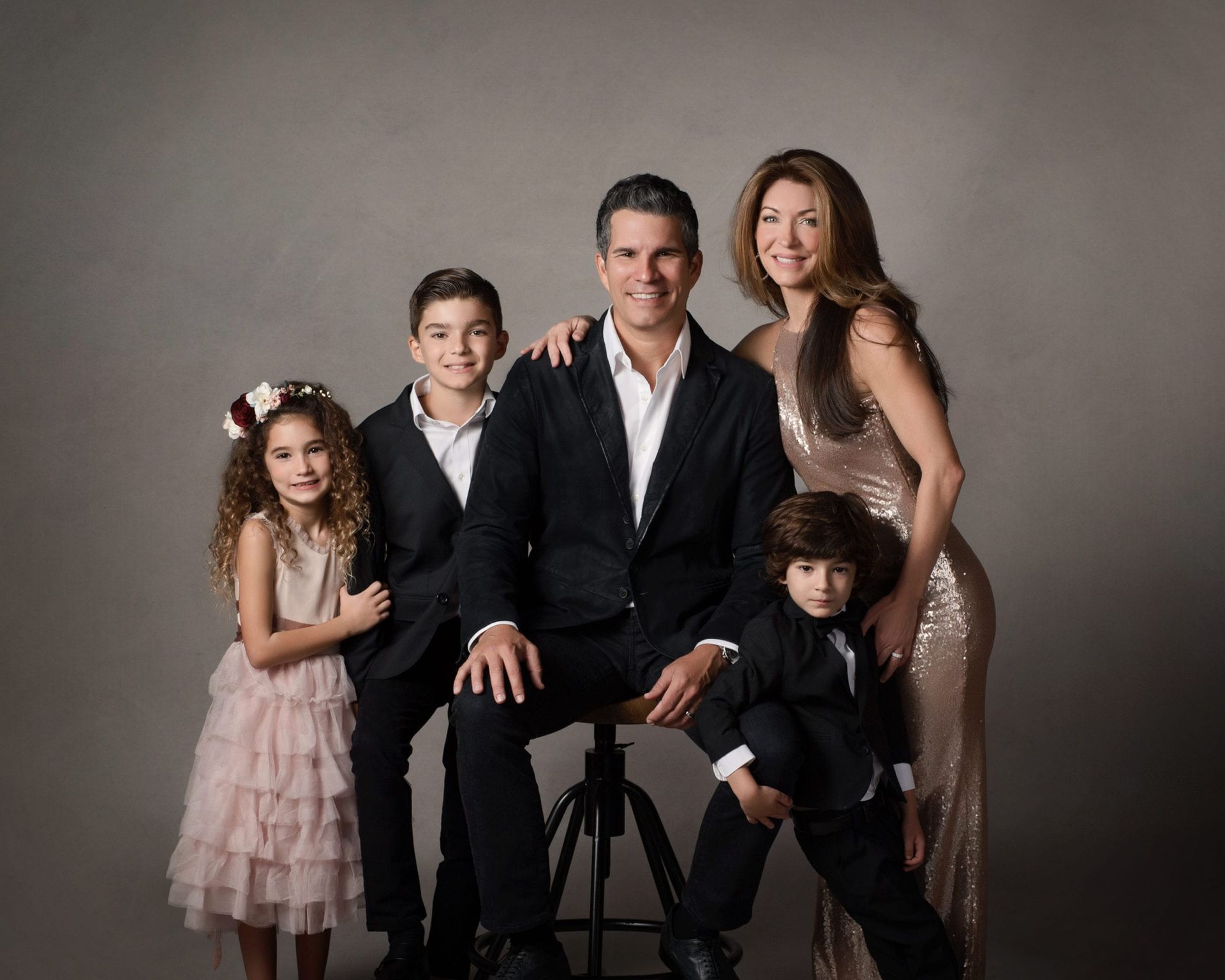 Family portrait: man seated, woman, and three children posing in formal attire. Gray background.