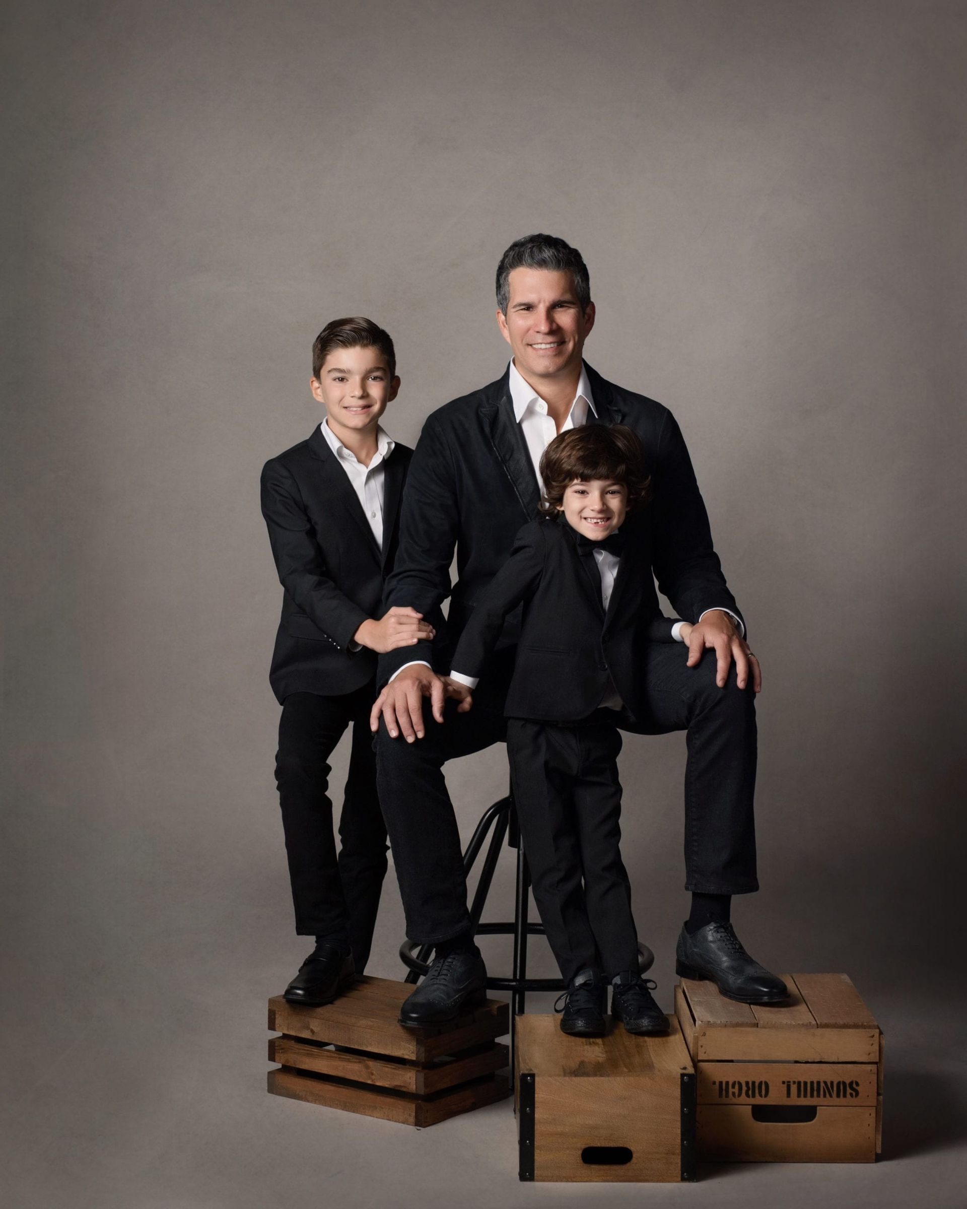Man and two sons pose in suits on boxes against a gray background.