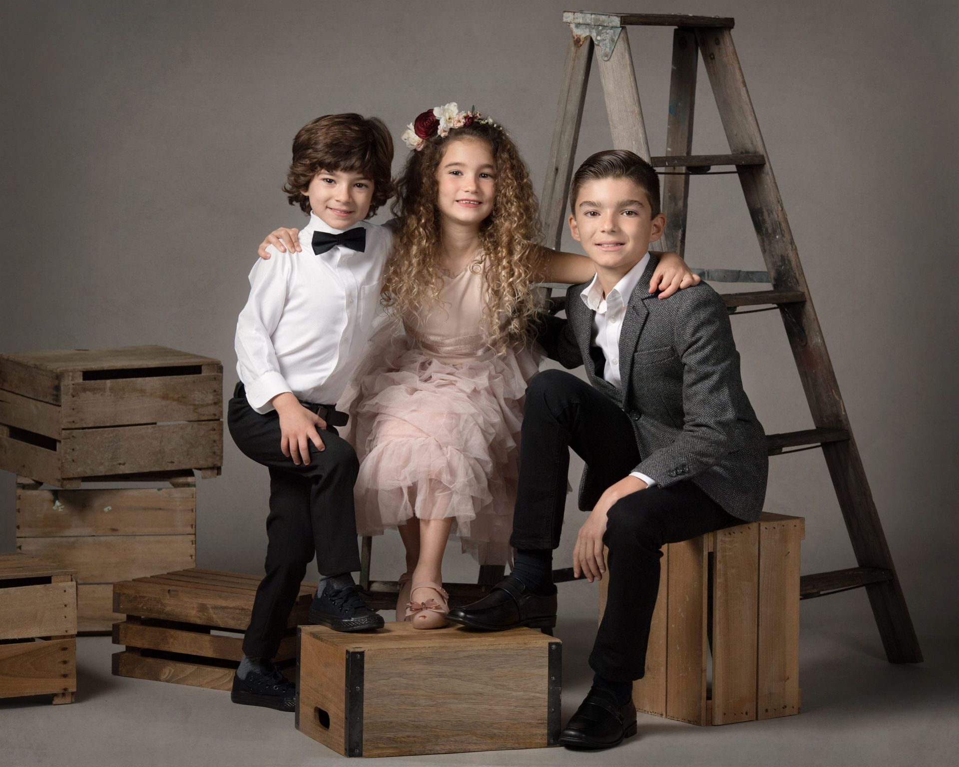 Three children pose; boy in tuxedo, girl in pink dress, and boy in blazer sit on wooden boxes by a ladder.