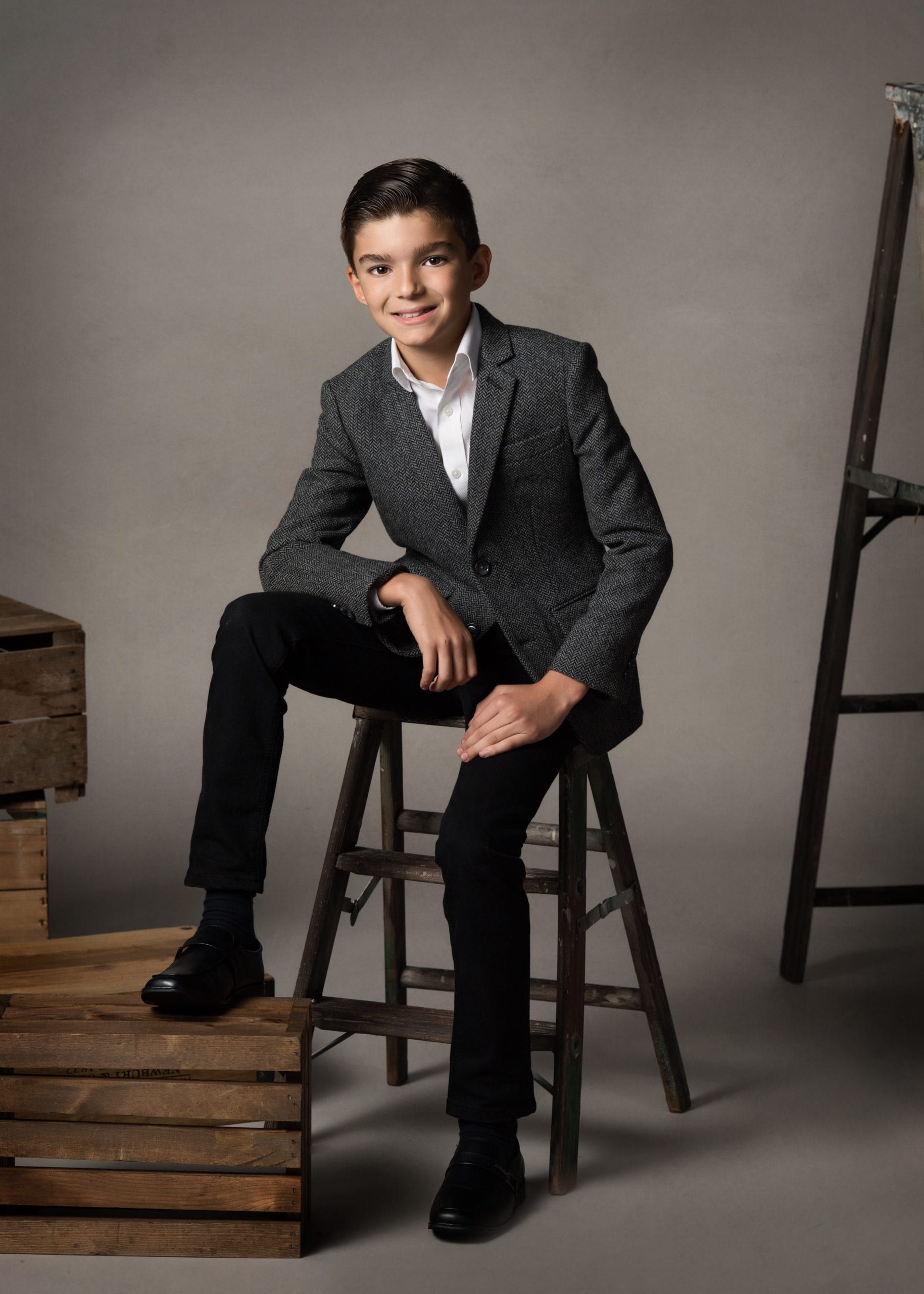 Teen boy in blazer, sitting on a stool, smiling. Neutral gray background, wooden crates, and a ladder.