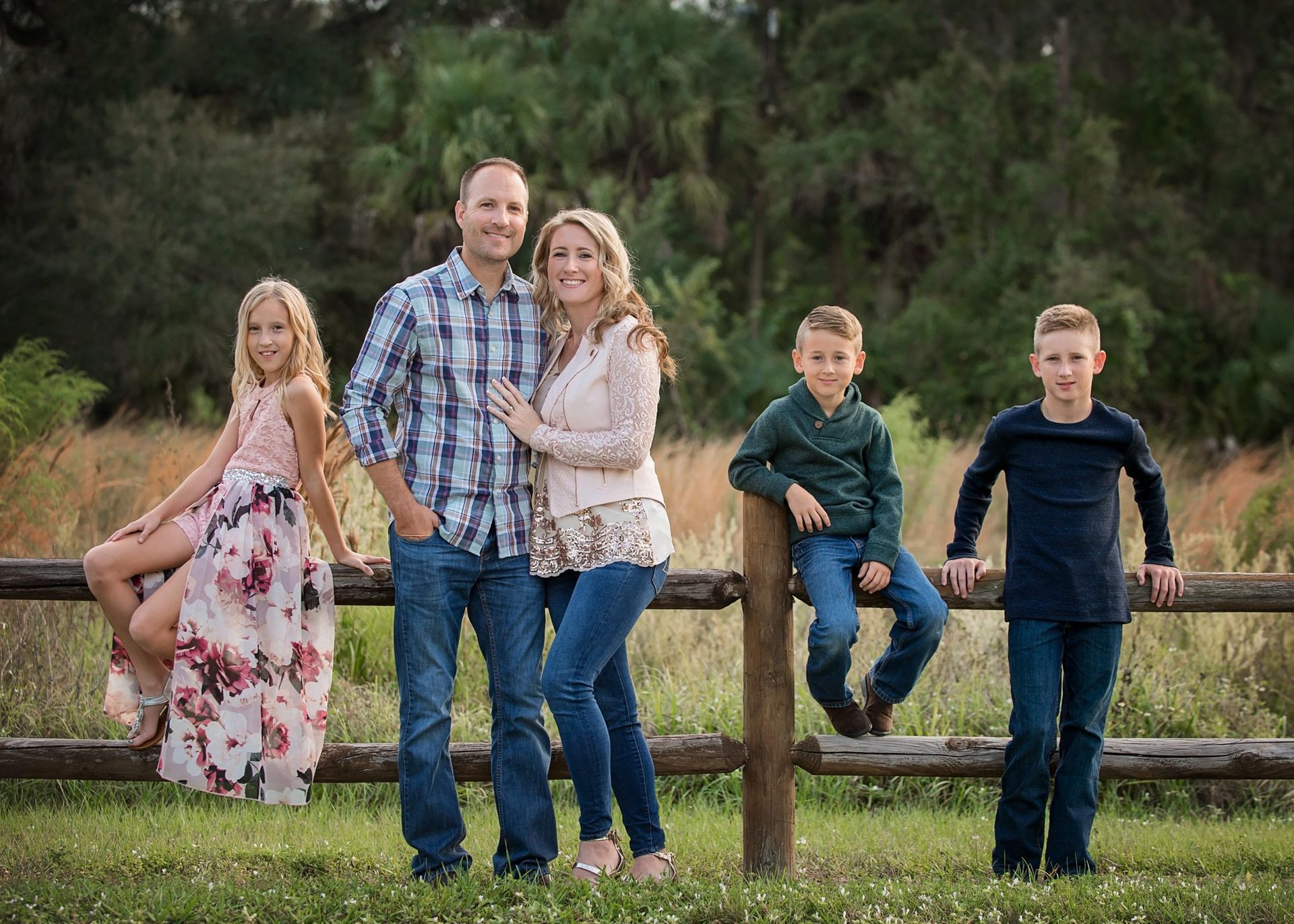 Family of five smiling outdoors near a wooden fence. Girl in floral dress, boys and parents in casual attire.