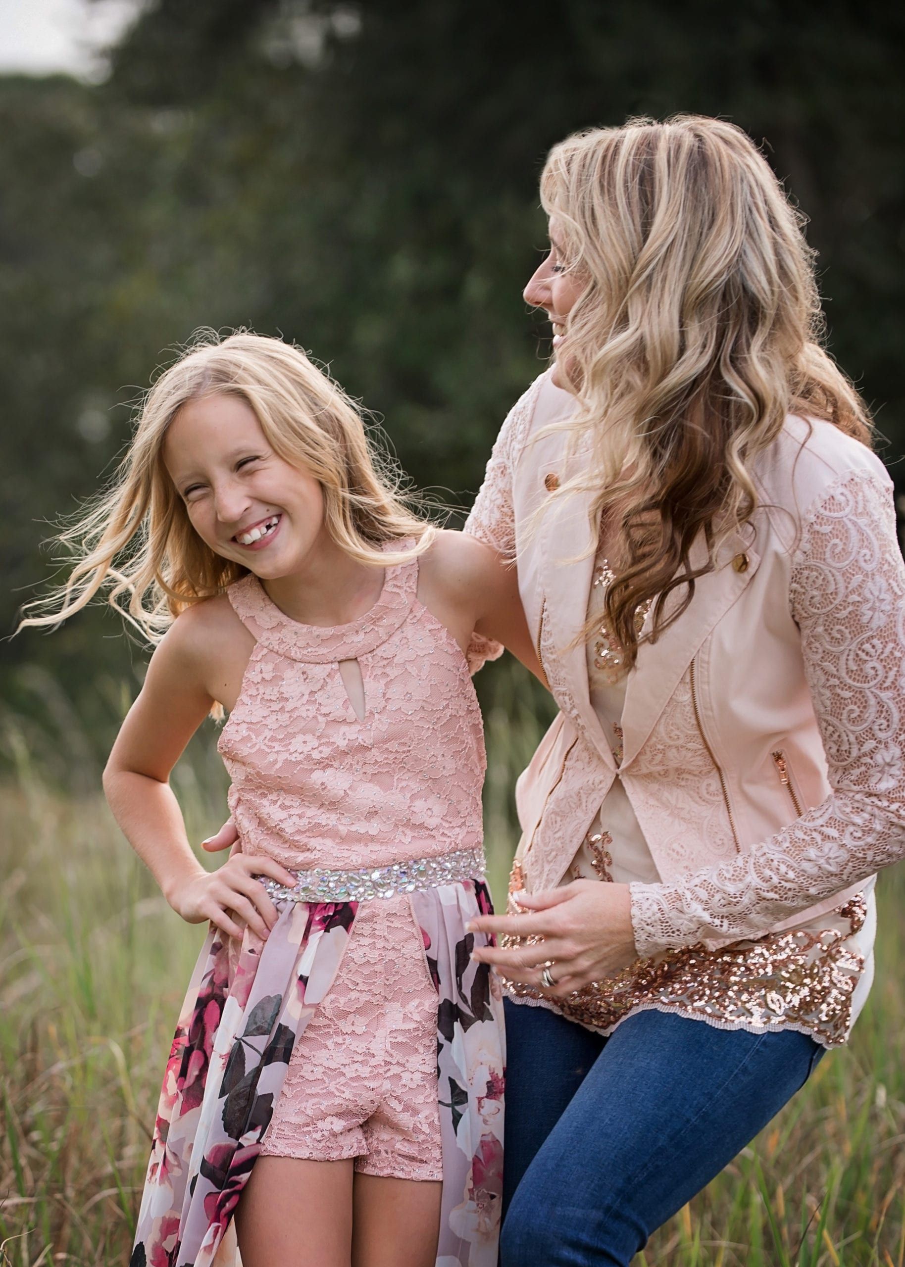 Woman and girl smiling outdoors; girl in pink lace outfit, woman in pink sequined jacket and jeans.