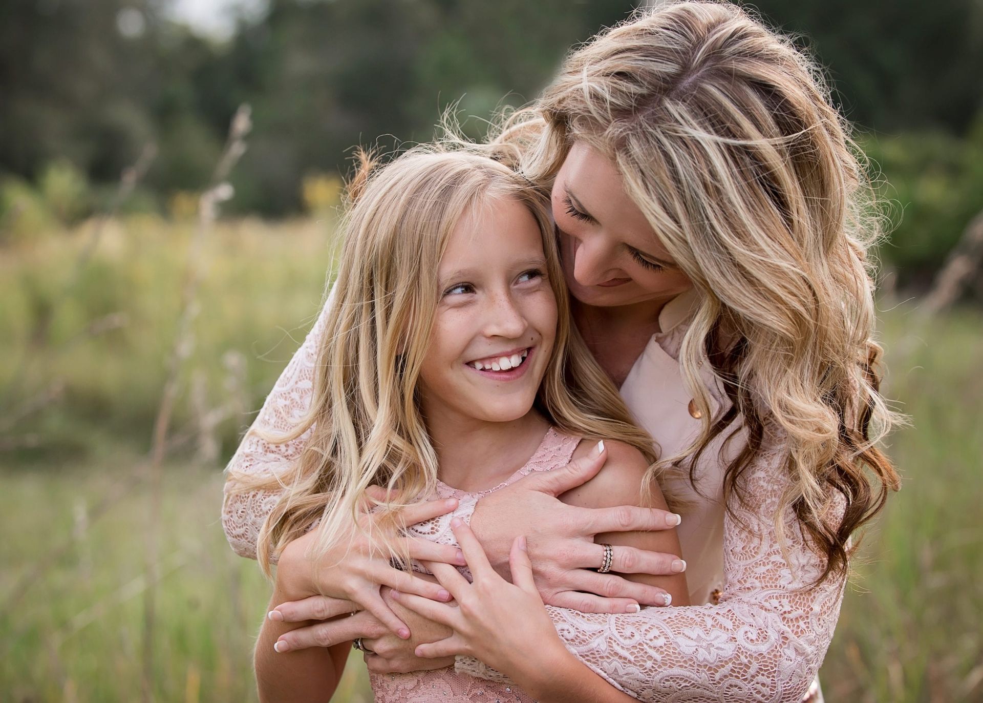 Mother embracing smiling daughter outdoors; both blonde, wearing pink dresses.