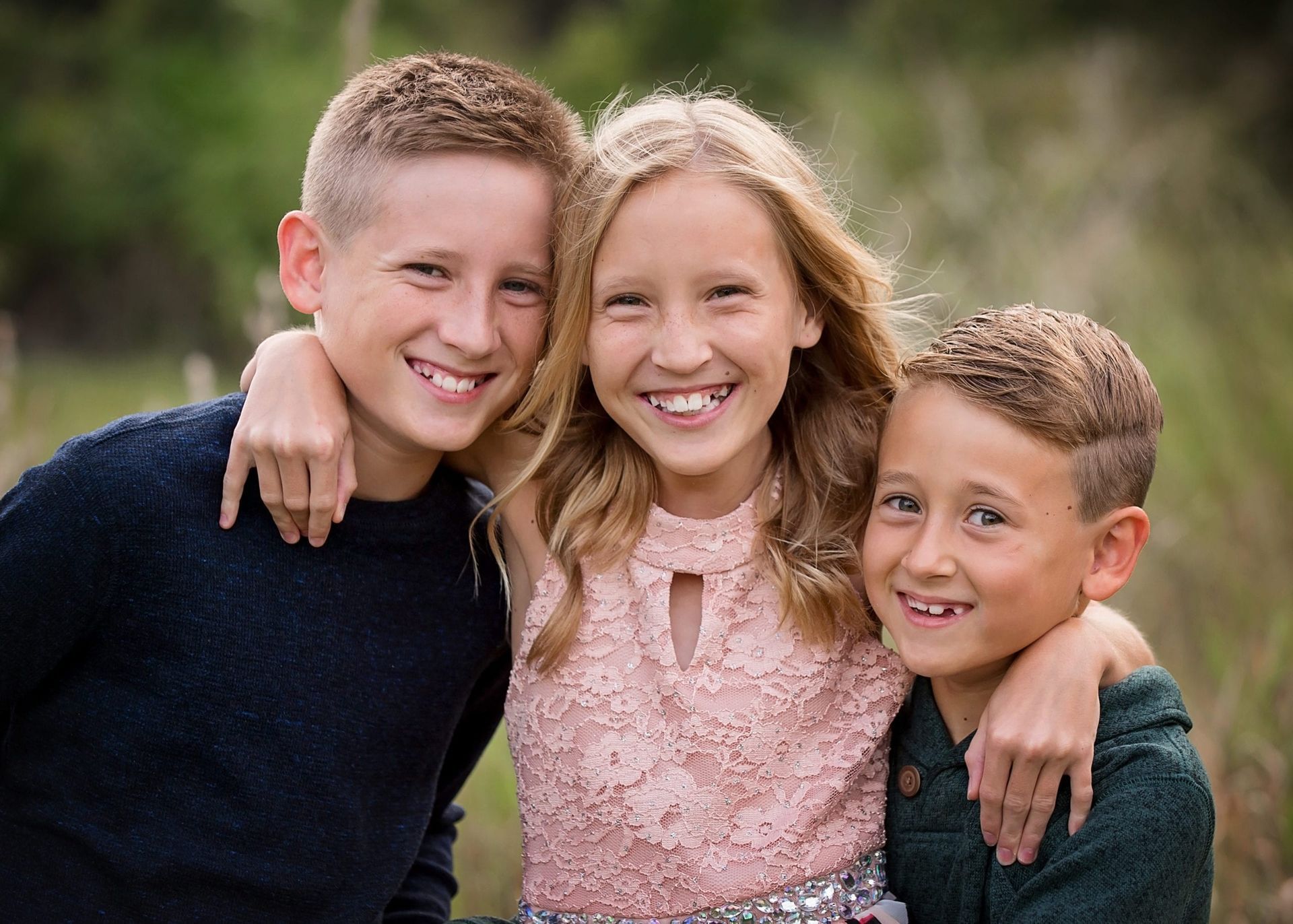 Three smiling kids, girl in the middle, arms around each other. Outdoor setting, natural light.