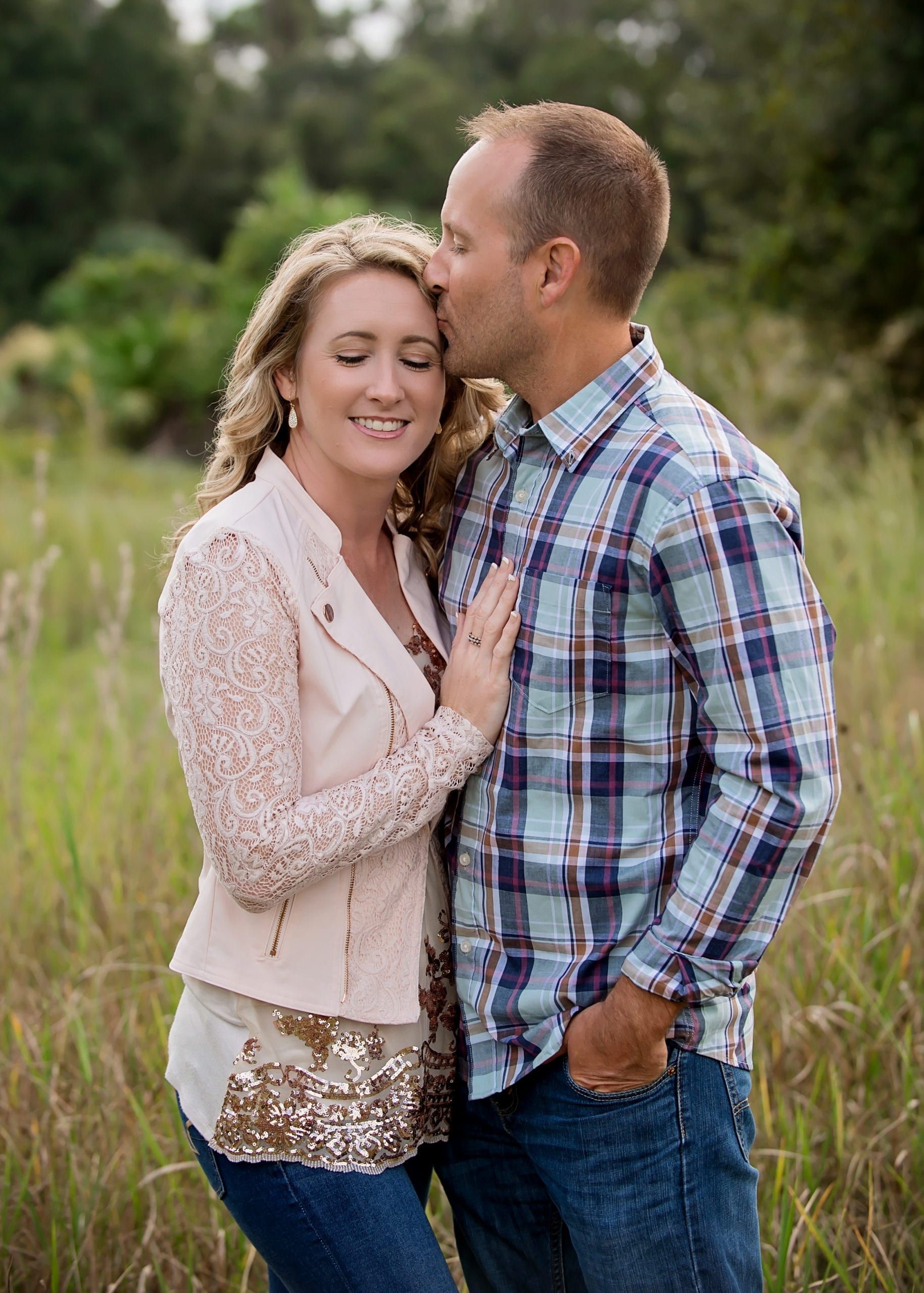 Couple embraces in grassy field, man kisses woman's forehead.