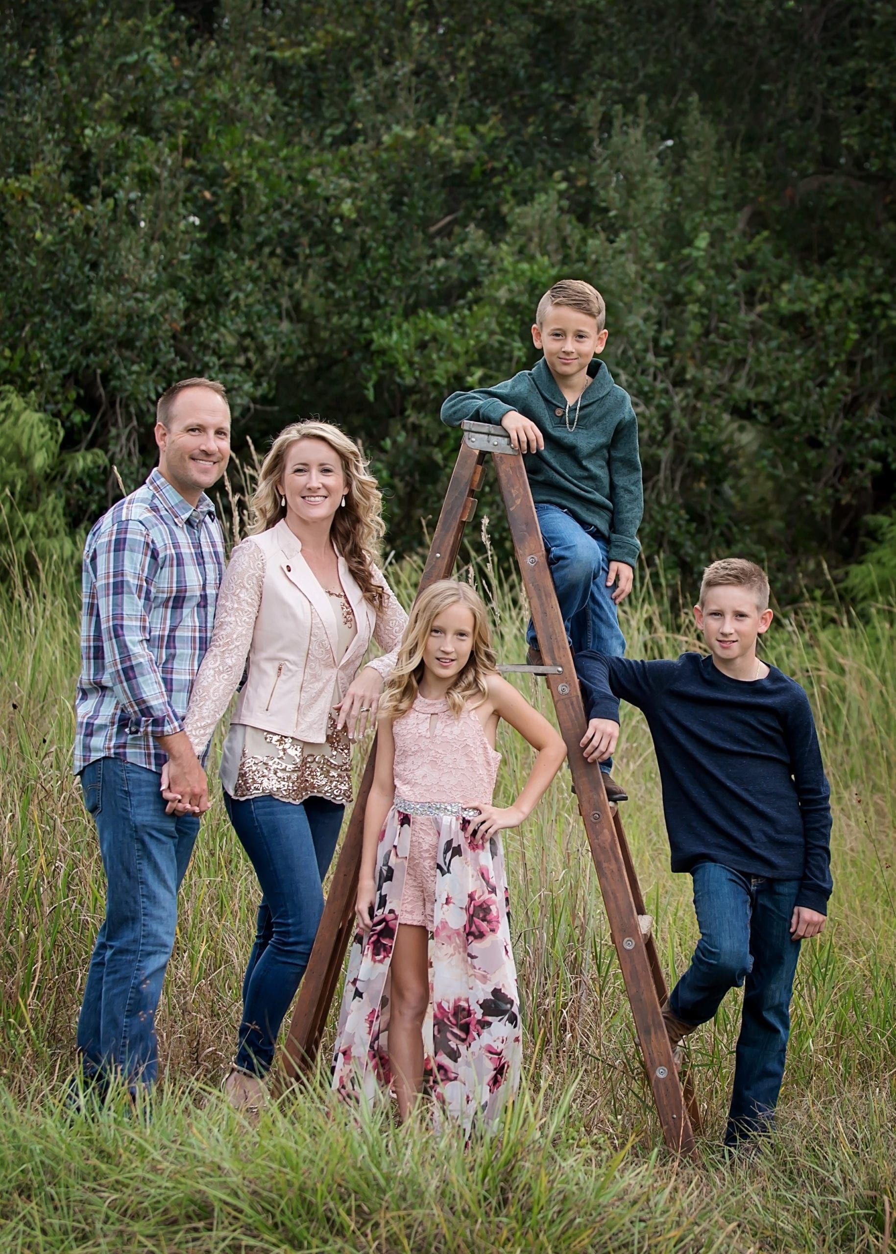 Family of five poses for photo in a field. Mom, dad, two sons, and daughter. Girl stands in front, boy on a ladder, other boy by the ladder.