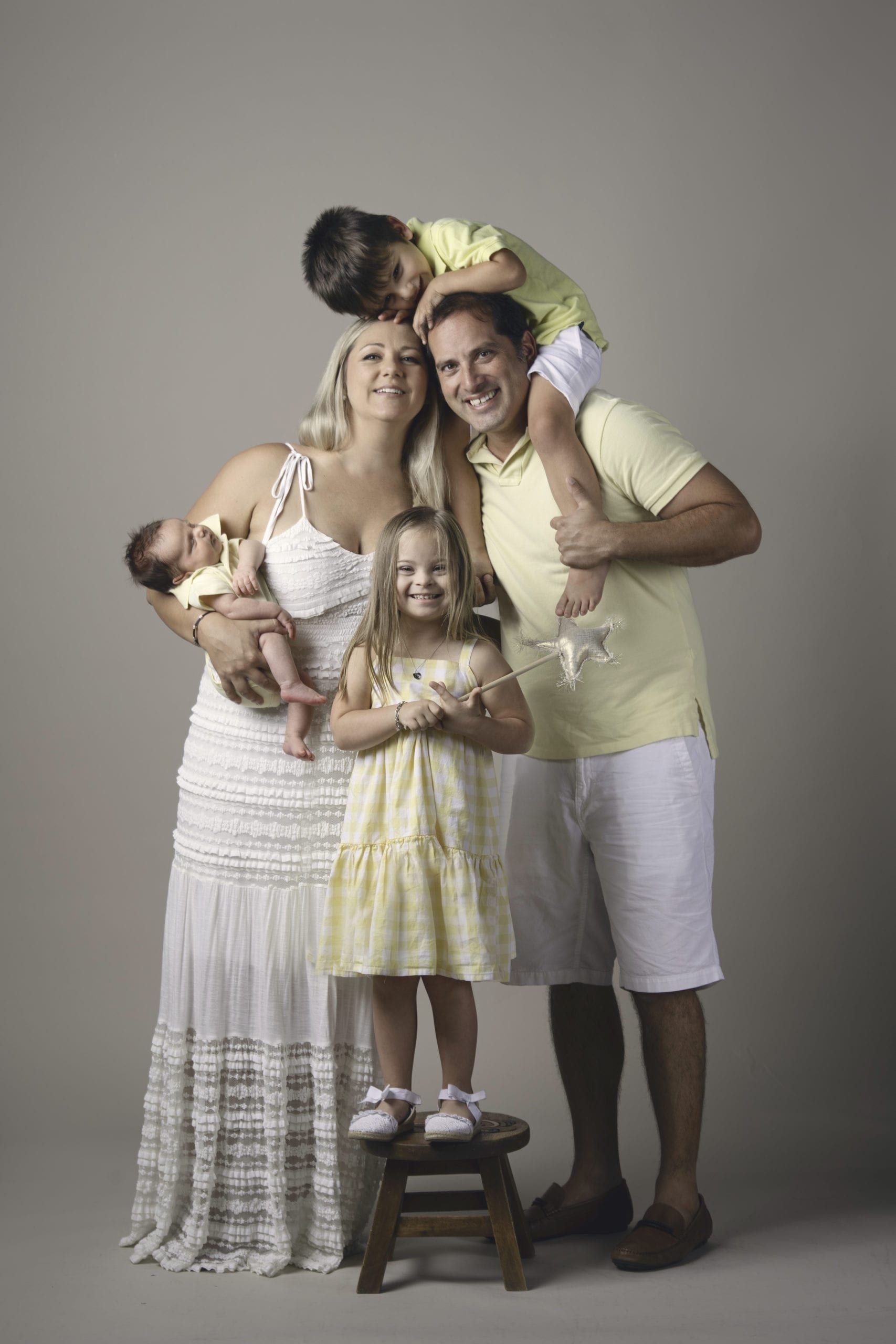 Family of five poses: parents, a baby, a child on a stool, and another on the father’s shoulders, all smiling.
