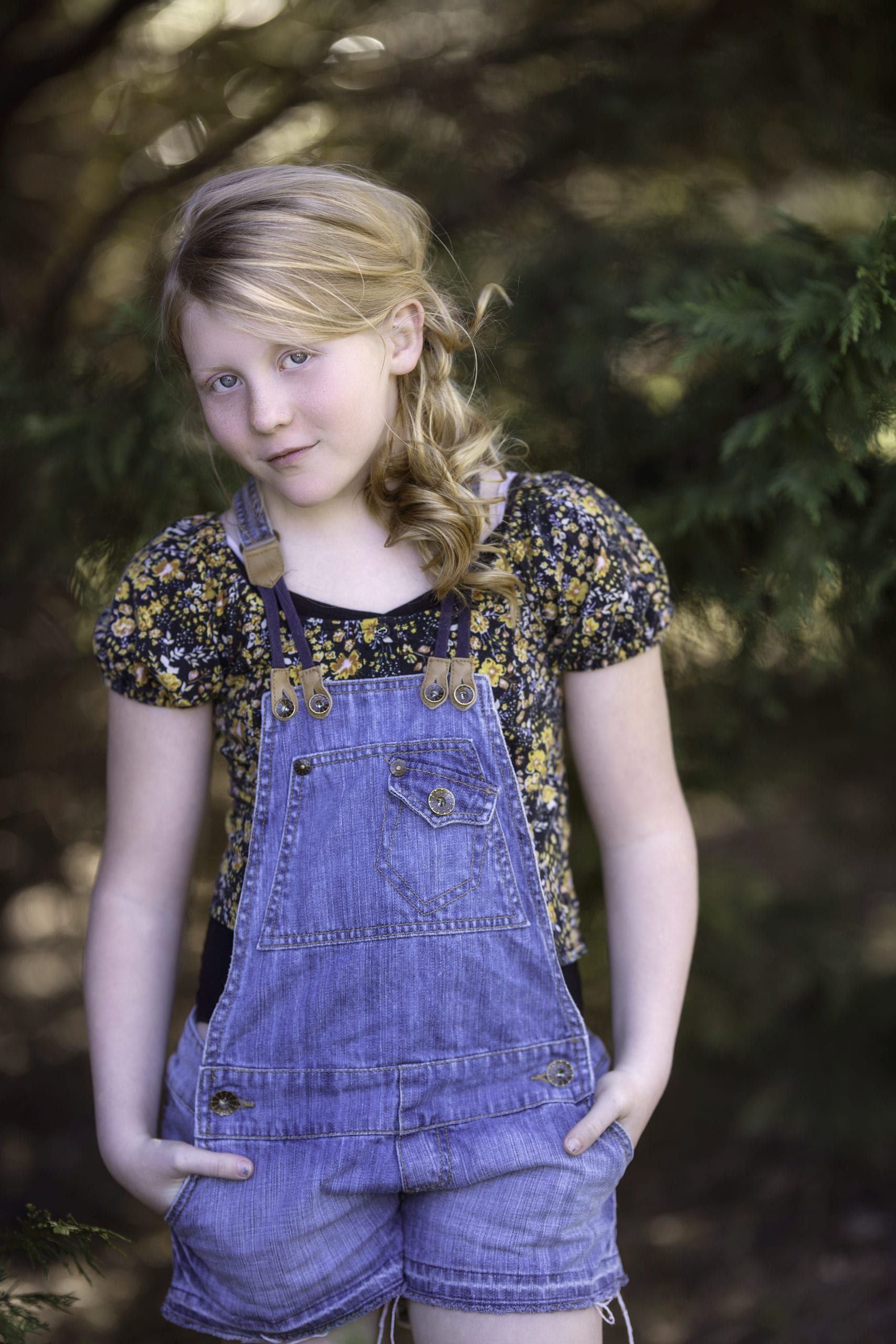 Young girl with blonde hair in denim overalls, hands in pockets, and floral top, outdoors.