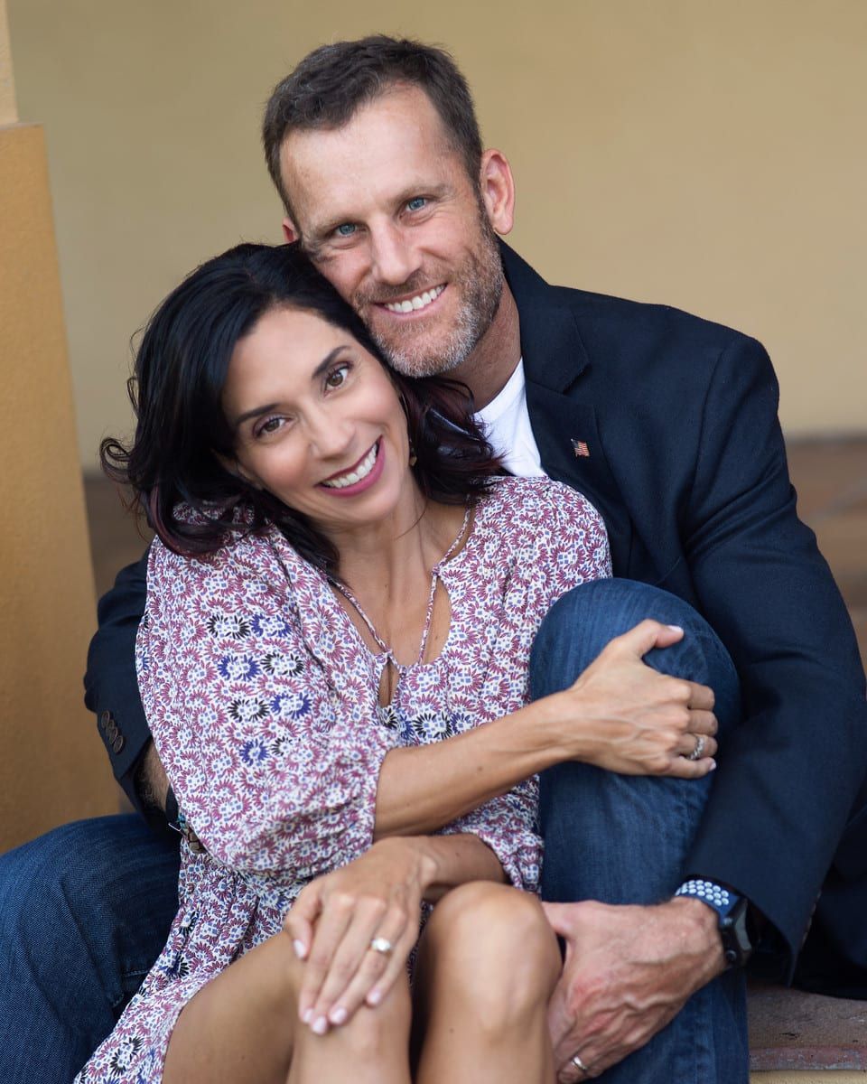 Couple smiling and embracing; woman in floral top, man in blazer, seated outdoors.
