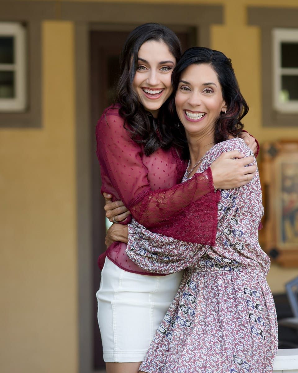 Woman hugs younger woman smiling outside; red top, white skirt, floral dress.