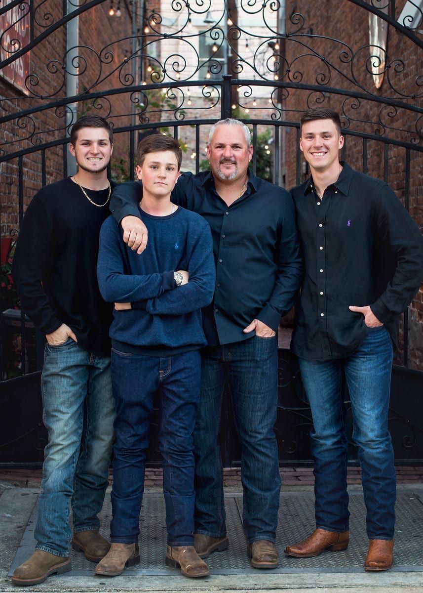 Family of four posing in front of a wrought iron gate. Men are wearing dark blue and jeans.