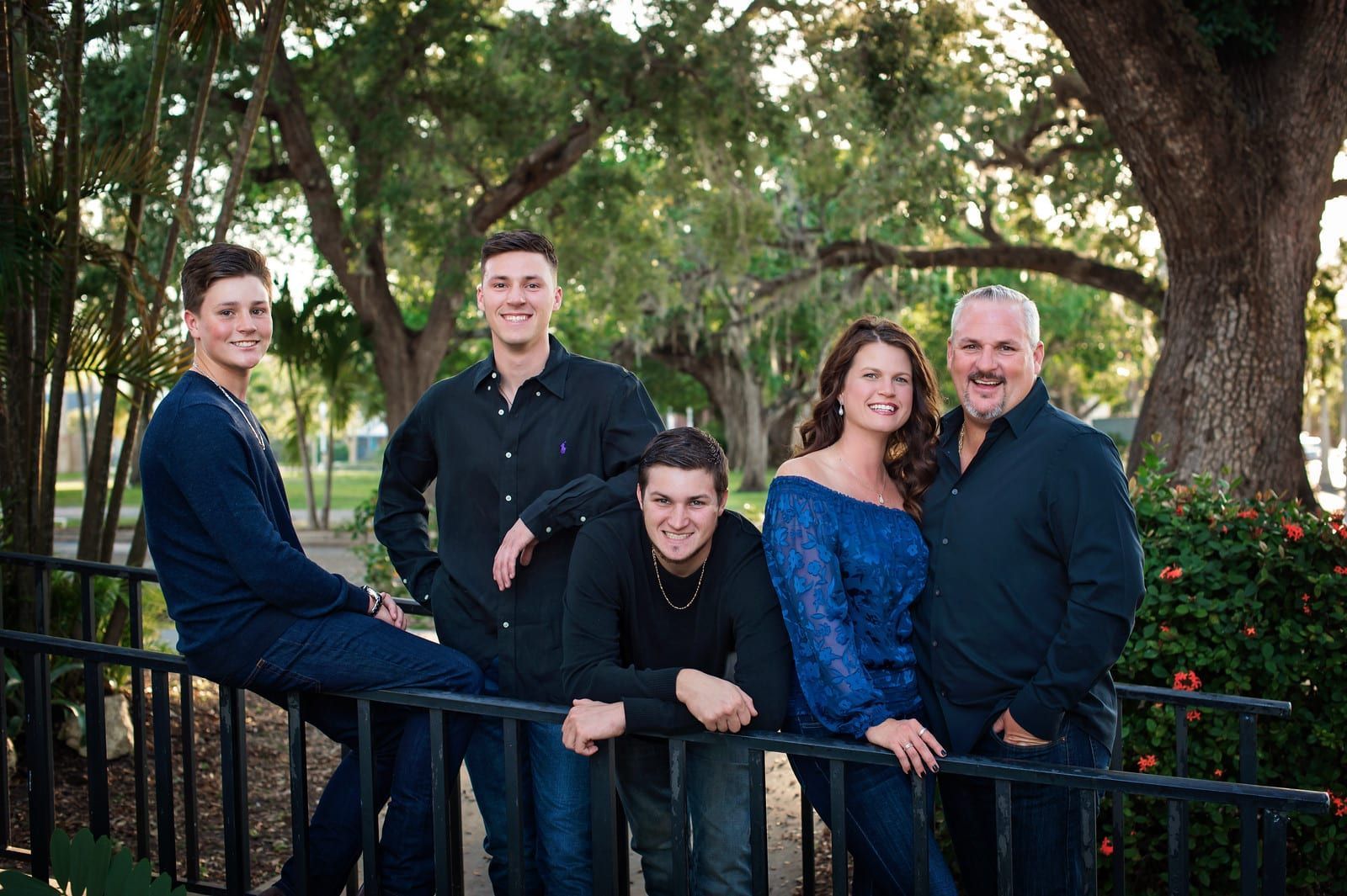 Family of five poses outdoors by a fence; smiles, greens, blues, and a happy vibe.