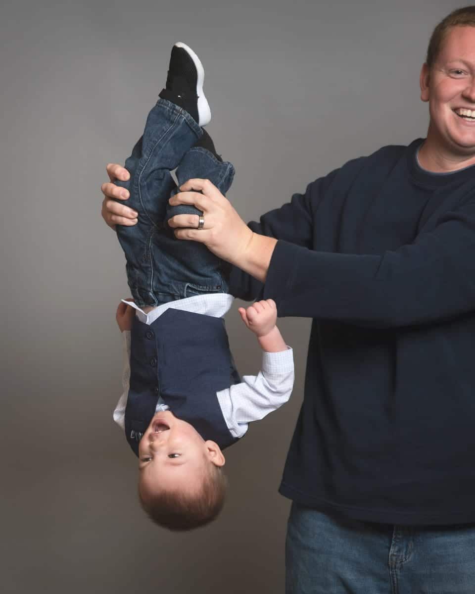 Man holding a baby upside down; both smiling against a gray backdrop.