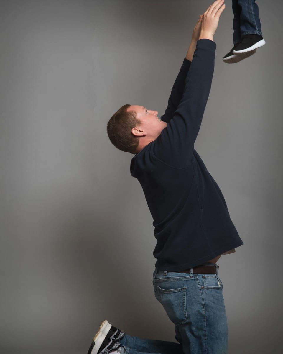 Man kneeling, holding child's legs up high, reaching arms up. Gray background, denim jeans.