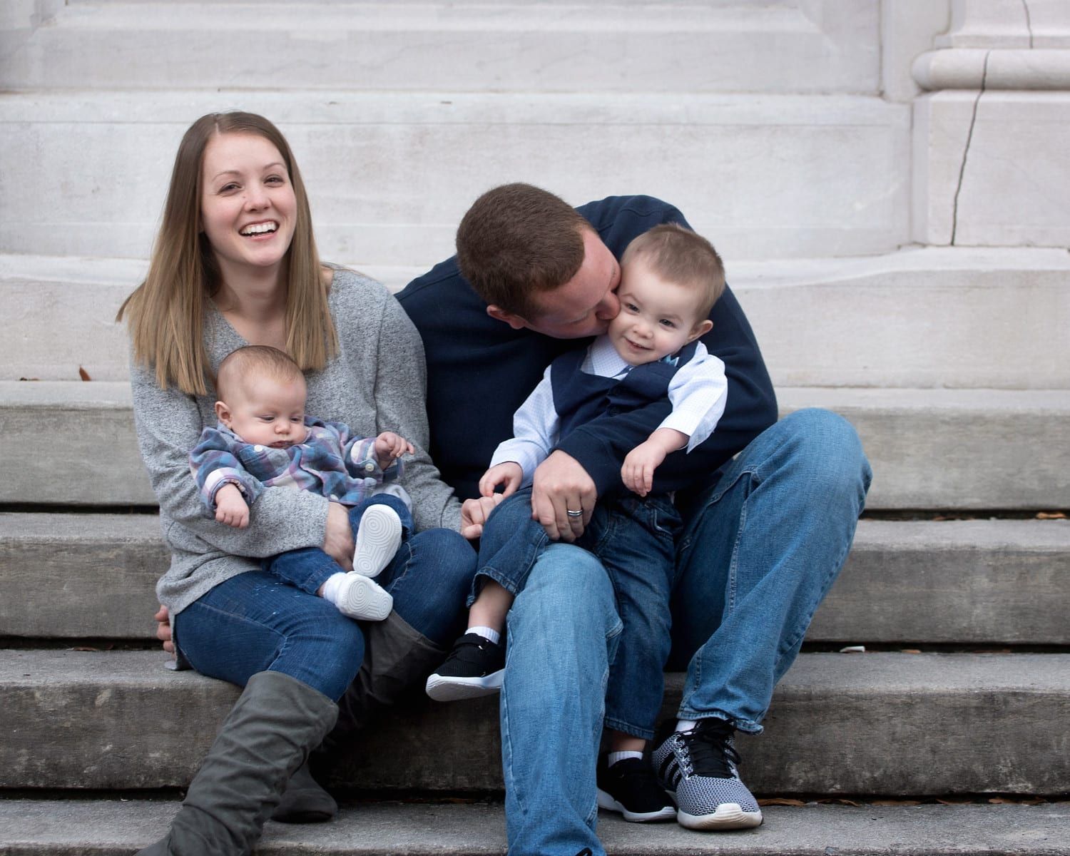 Family of four sits on steps, mom smiles holding baby, dad kisses toddler.