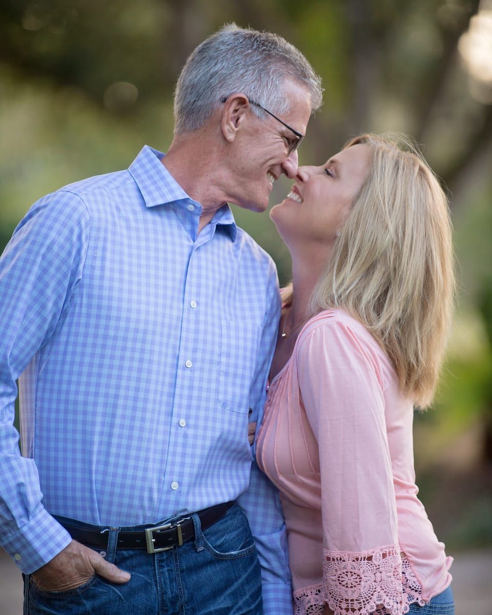 A smiling couple embraces outdoors. The man wears blue, the woman, pink. They appear happy.