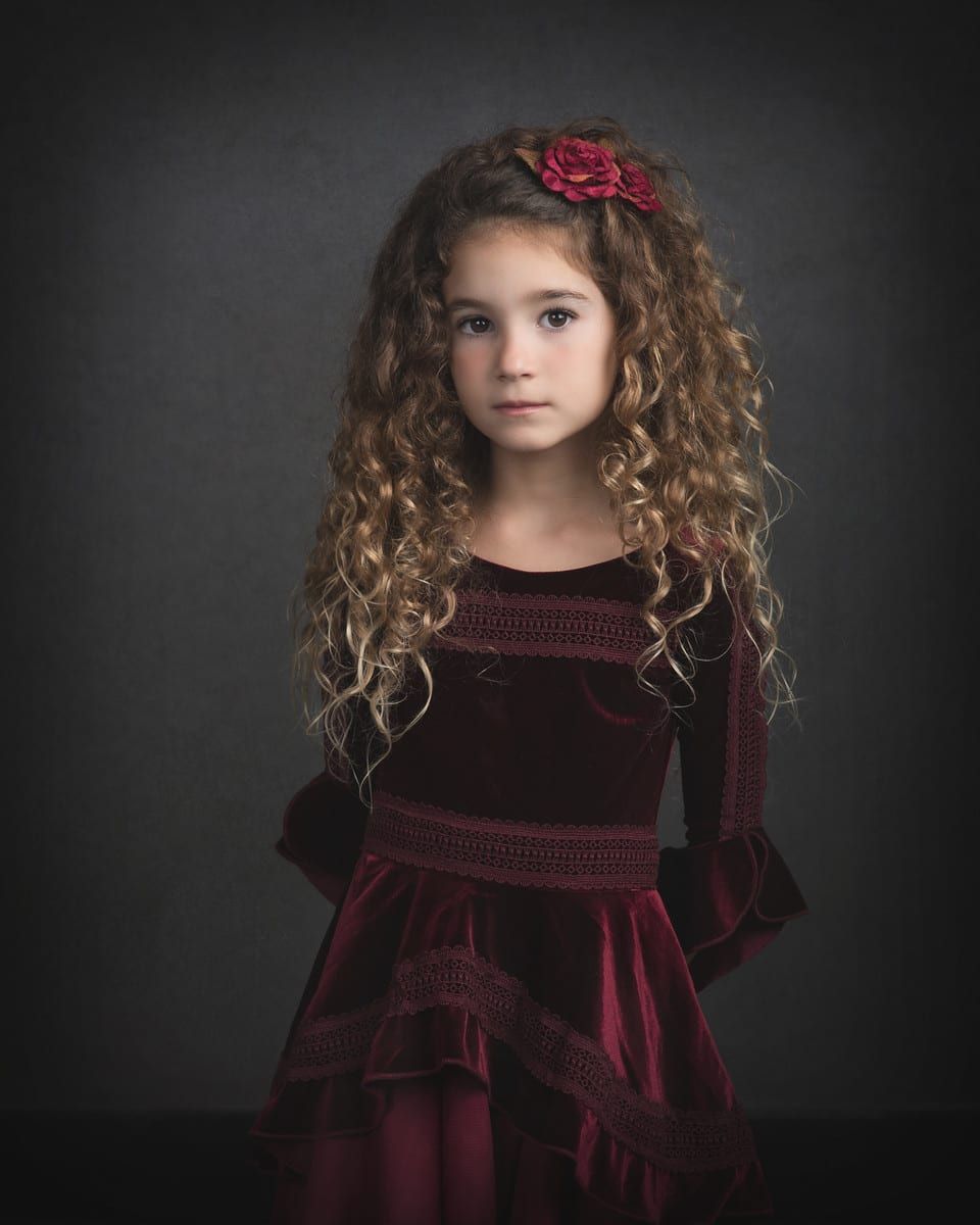 Girl with curly brown hair in a burgundy velvet dress and a flower hair clip, hands behind her back, against a dark background.