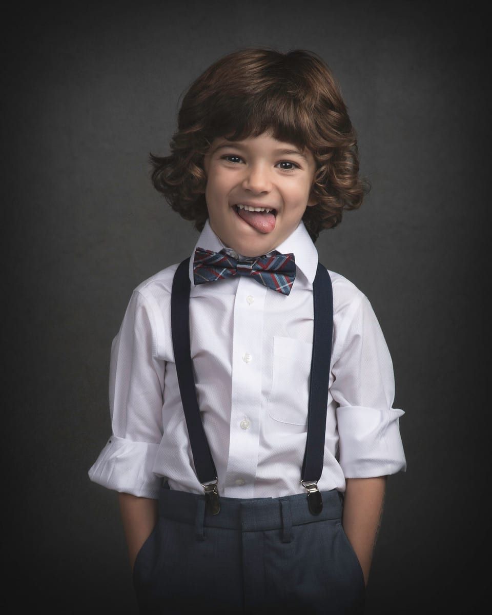 Boy with curly brown hair, wearing a bow tie, suspenders, and white shirt, sticking his tongue out.