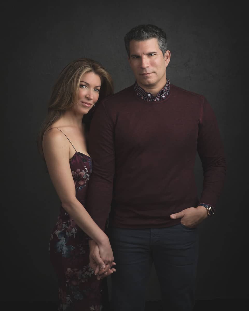 A couple poses formally against a dark backdrop. The woman leans on the man, holding hands. Both have serious expressions.