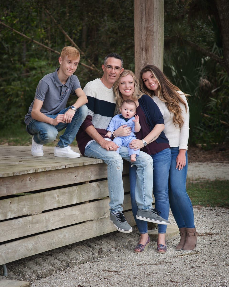 Family of five poses for a photo outdoors: two teens, parents, and a baby.