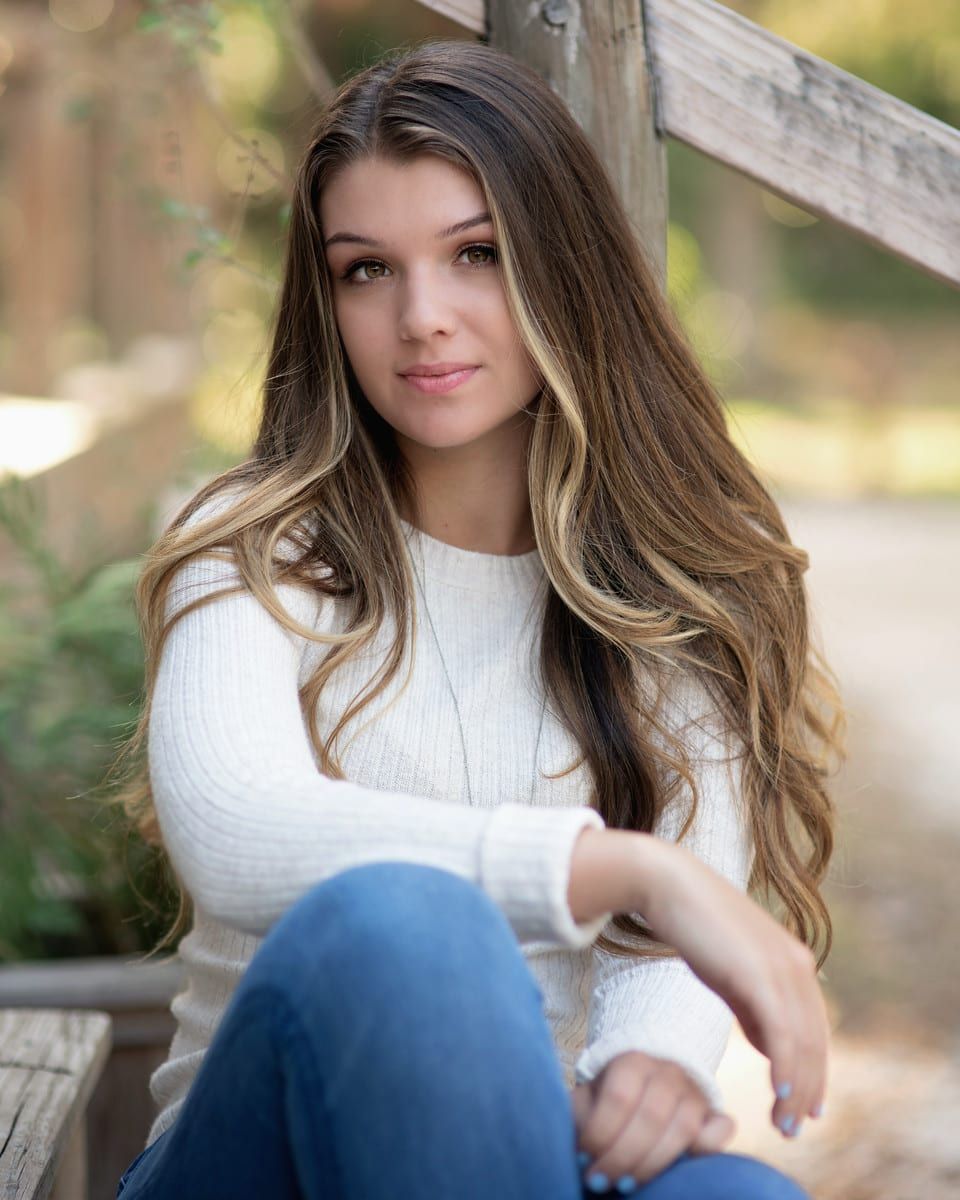 Young woman with long brown hair and highlights, wearing a white sweater and blue jeans, outdoors.