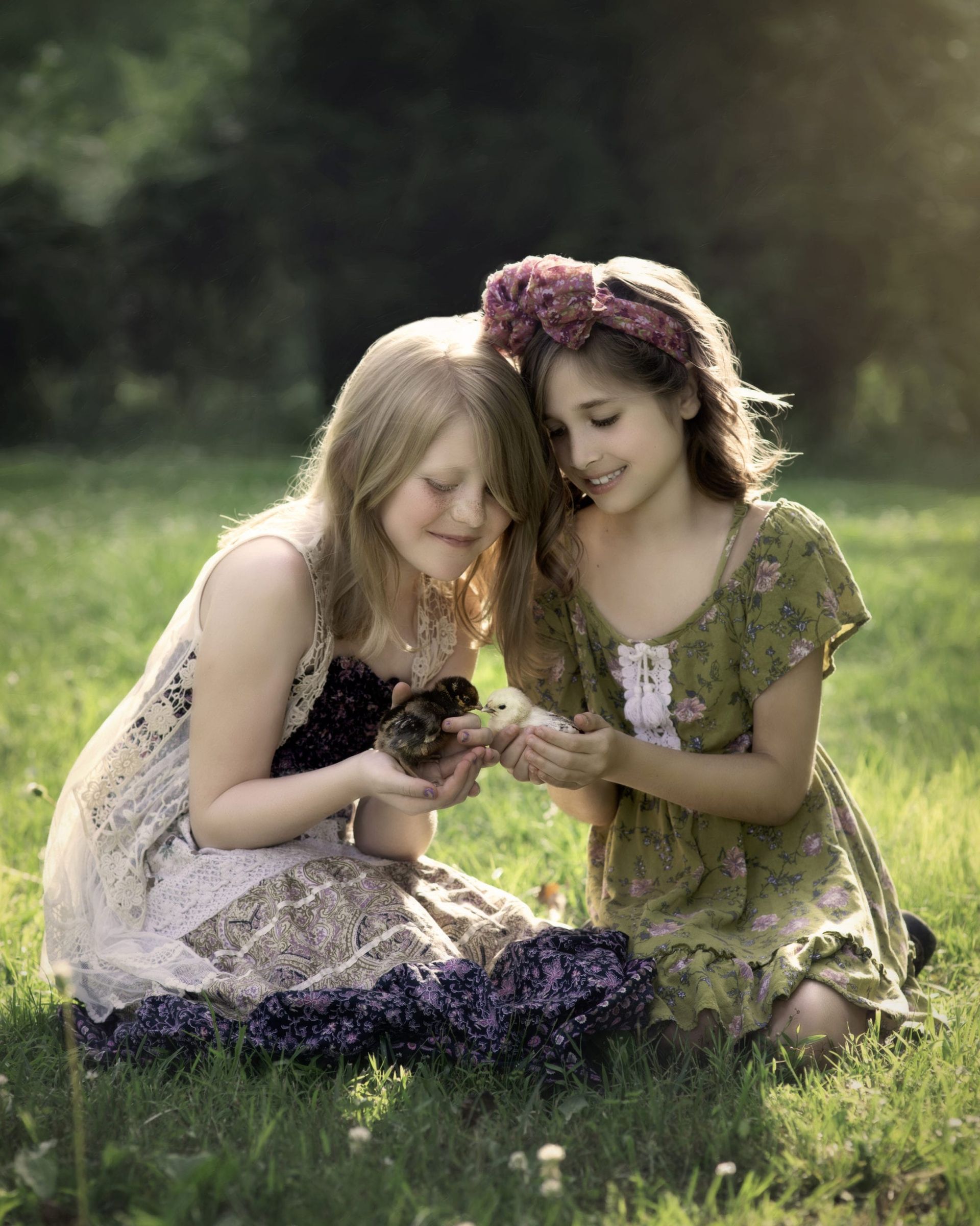 Two girls in dresses on grass, holding a small bird, smiling.