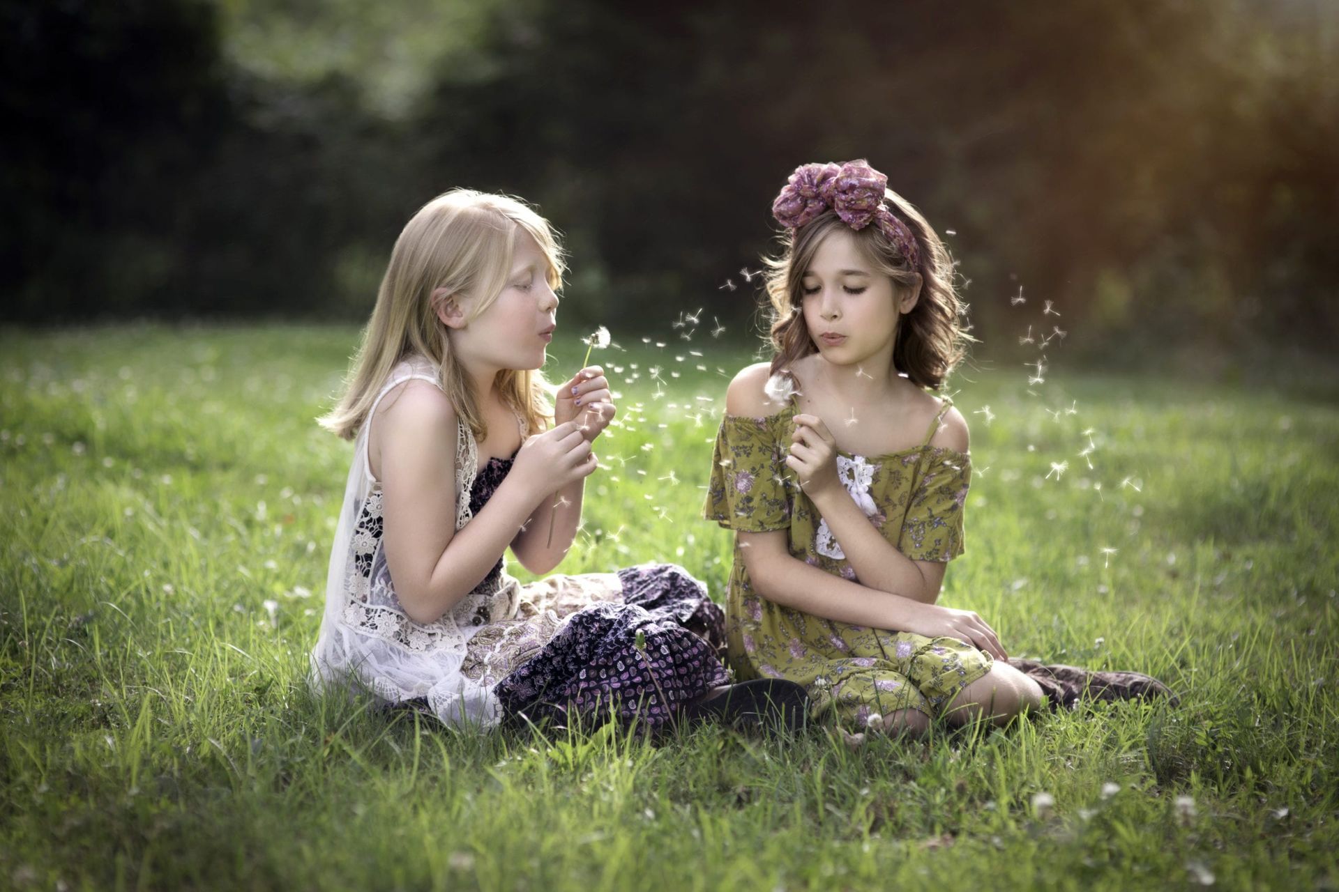 Two young girls blowing dandelion seeds in a grassy field; one wears a flower crown.