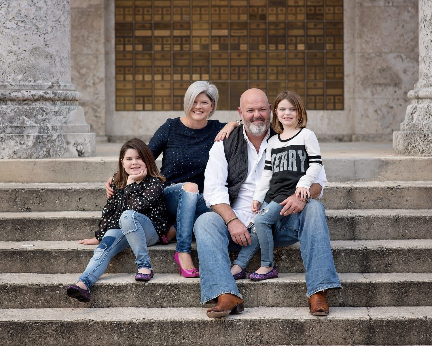 Family of four smiling, posing on stone steps; parents in the middle, two daughters on each side.