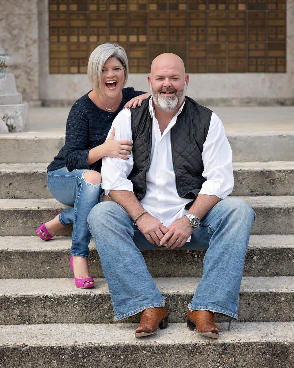 Woman with gray hair and man with beard sitting on steps, smiling; stone backdrop.