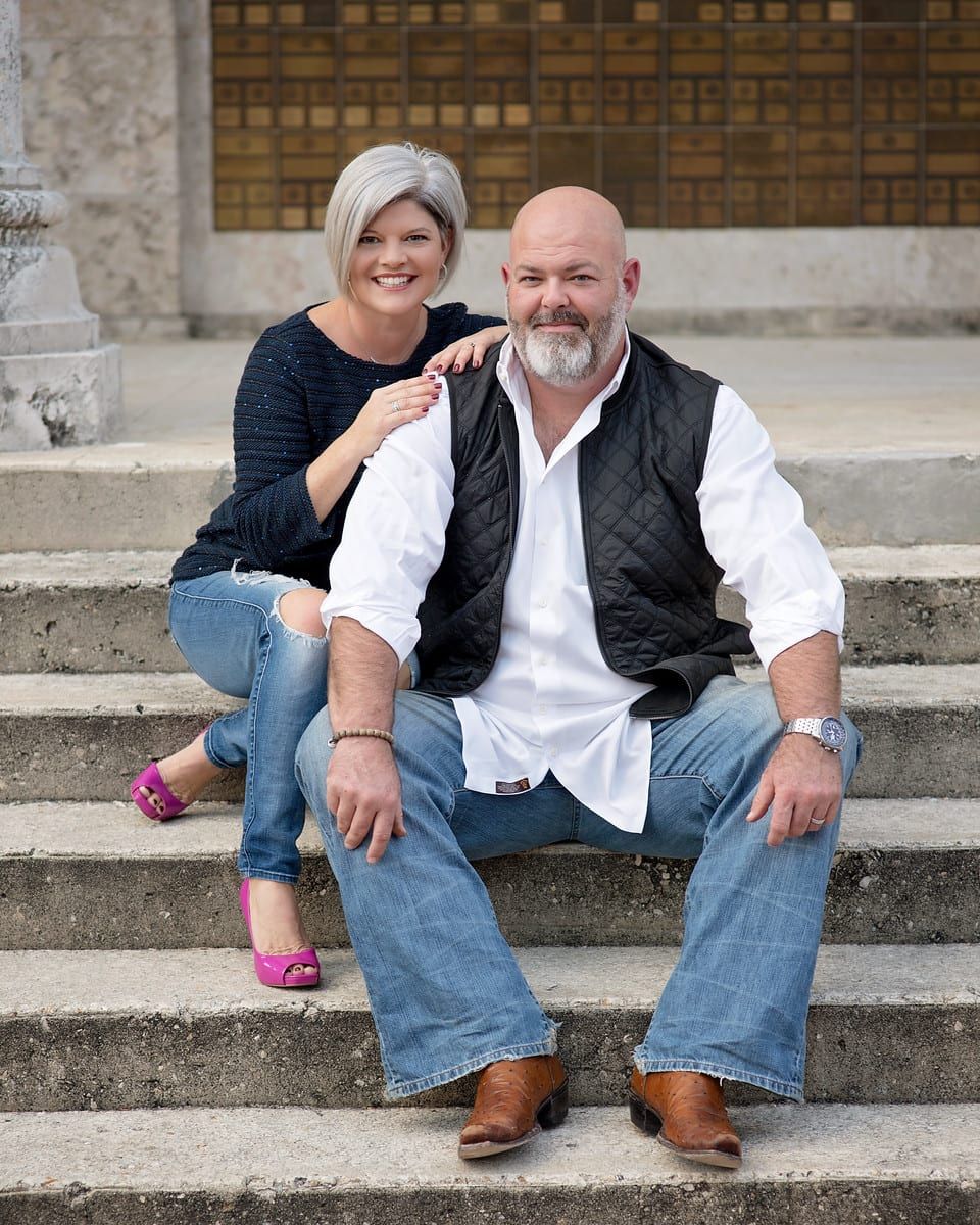 Couple sits on steps; woman in blue, pink shoes, man in black vest, white shirt, and jeans.