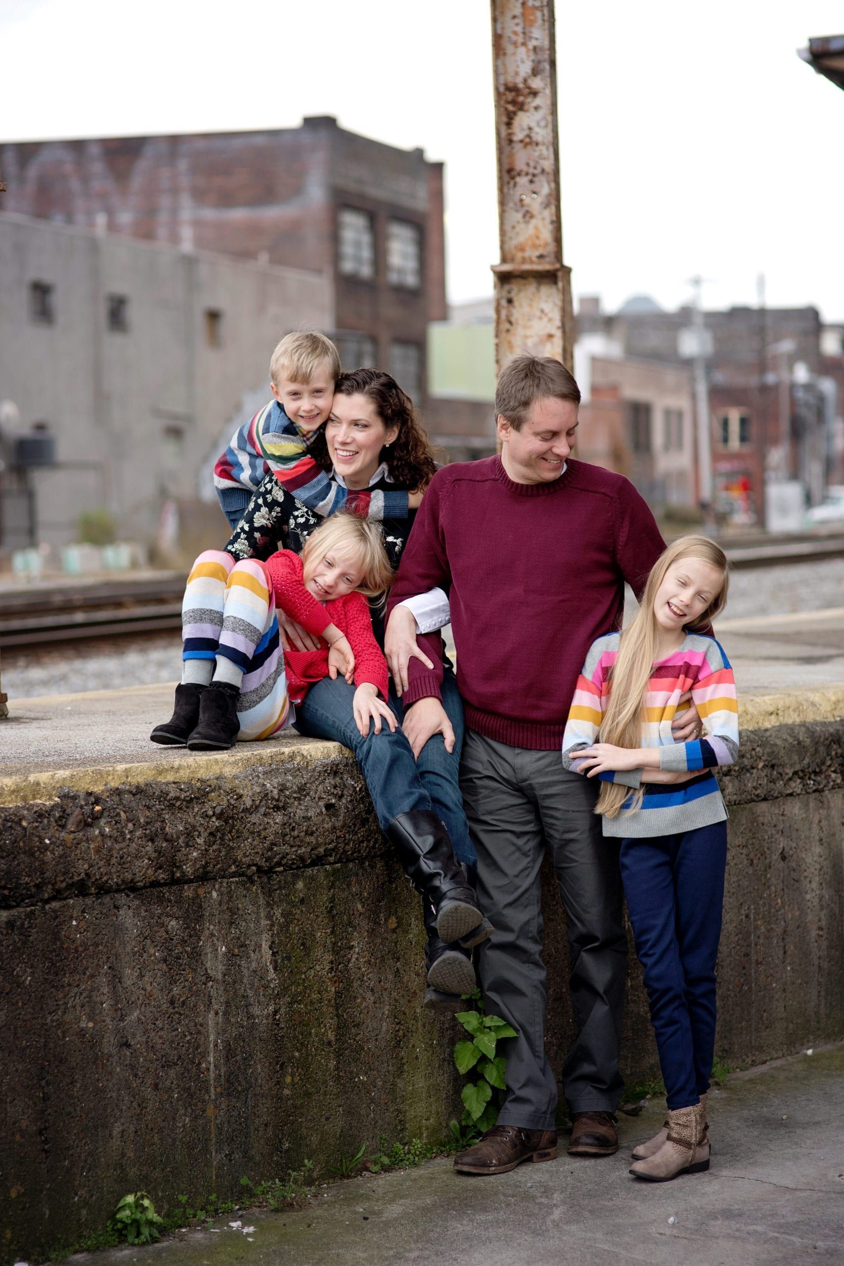 Family of five poses together on a concrete ledge near train tracks; buildings in background.