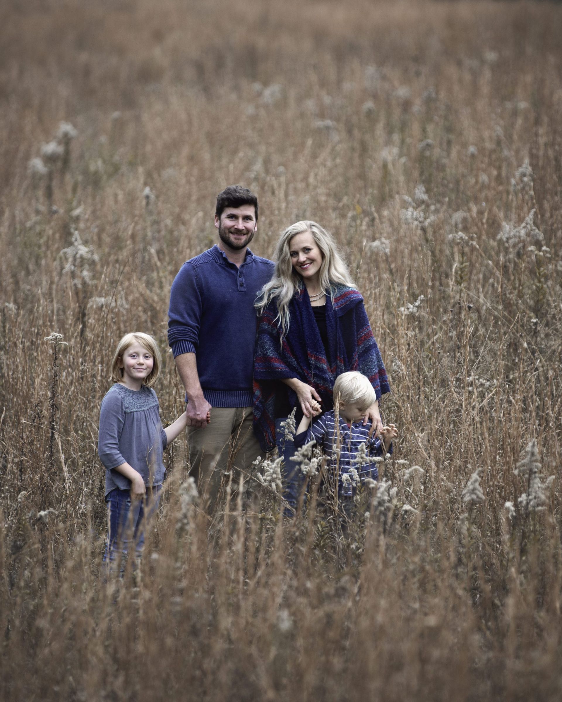 Family of four standing in a field of tall grass, smiling. Parents with young children; autumn colors.