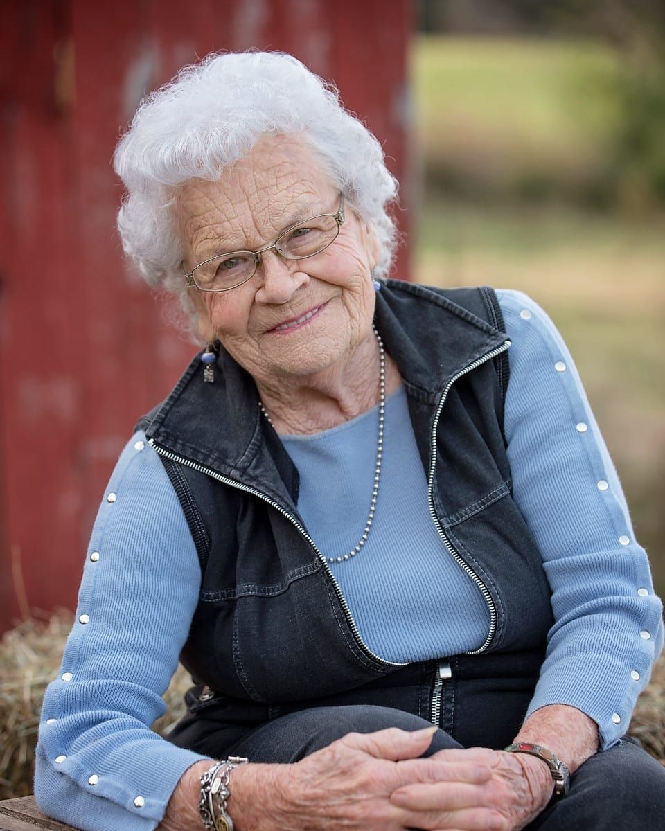Elderly woman with white hair and glasses smiles, wearing blue and black vest, sitting outdoors.