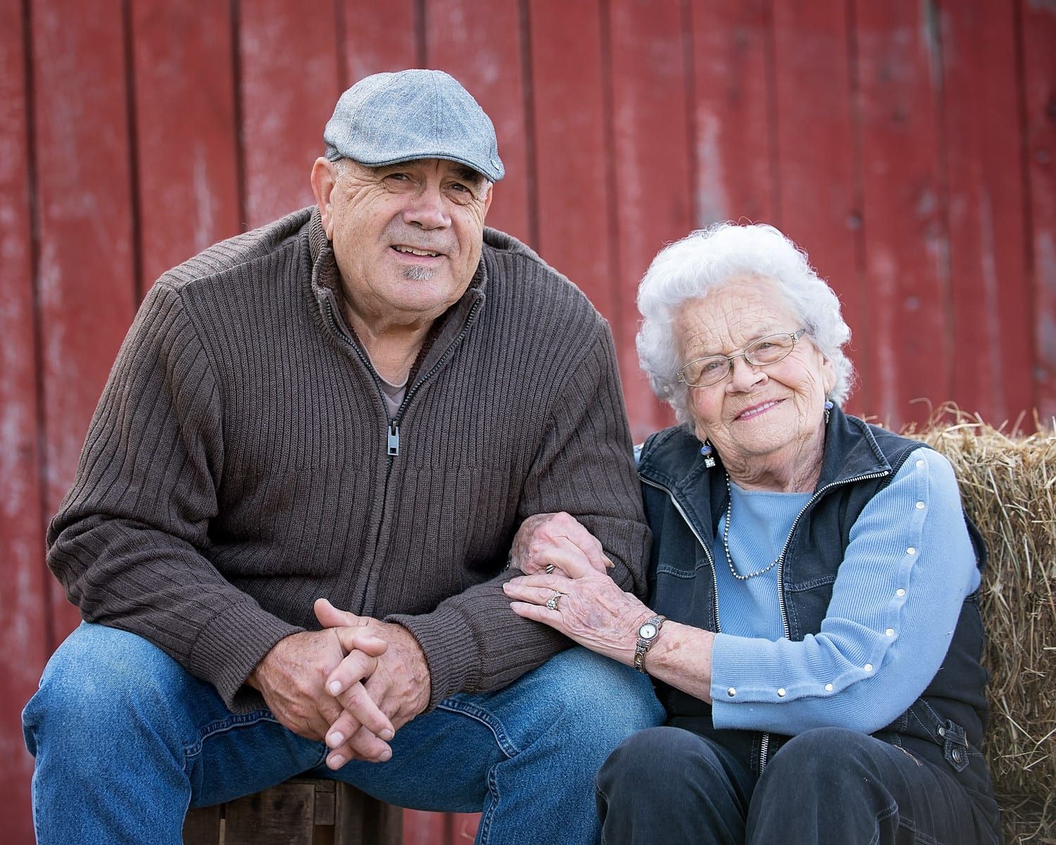 Elderly couple sits together outdoors. Man in cap and sweater, woman in glasses and vest. Red barn backdrop.