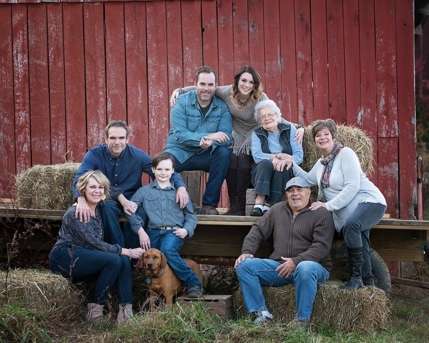 Family poses in front of a red barn; includes several adults, a child, a dog, and hay bales.