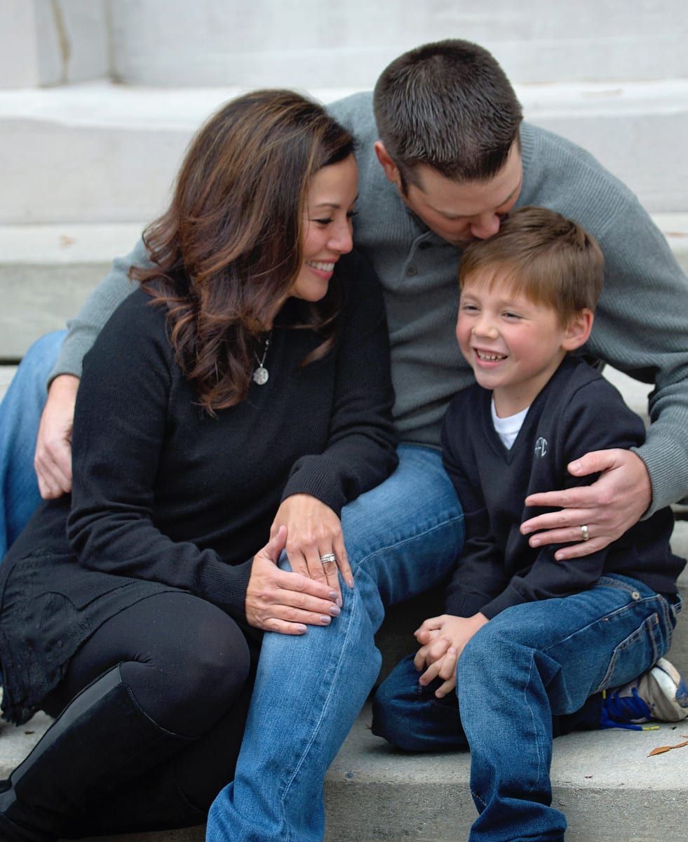 Family of three smiling on outdoor steps. Dad kisses son's head. Mom looks at son.