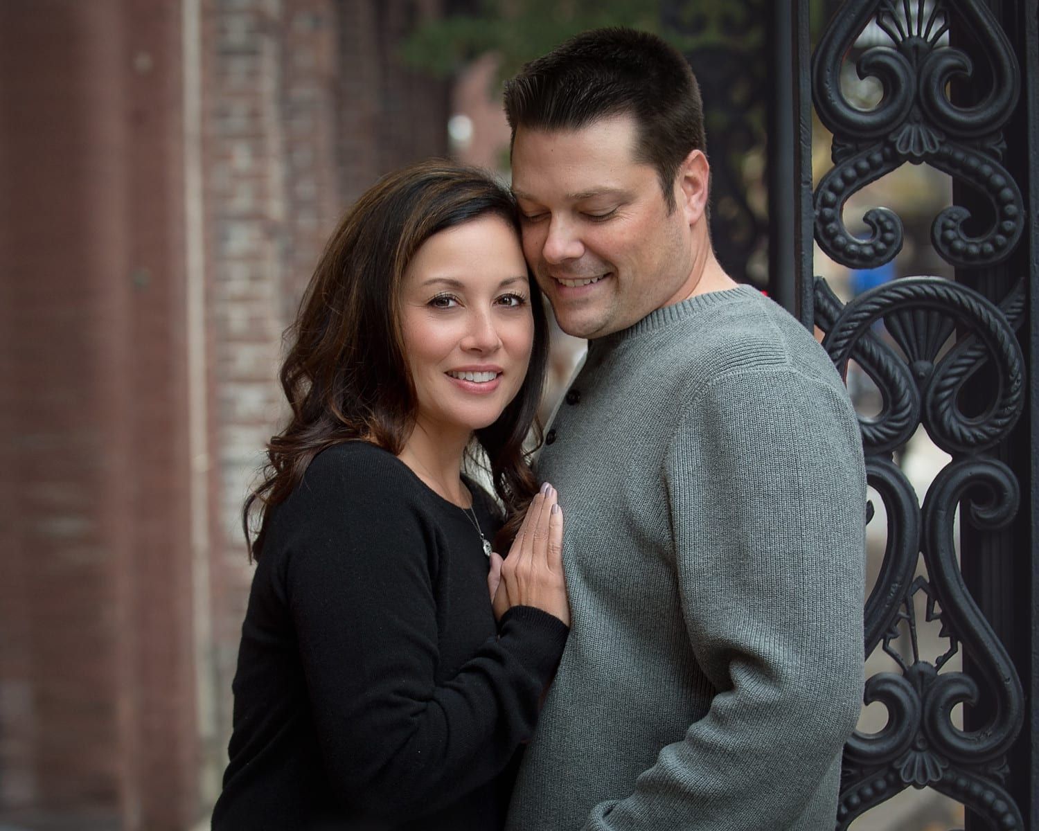 Couple embraces near an ornate gate; woman in black top, man in grey sweater, smiling.
