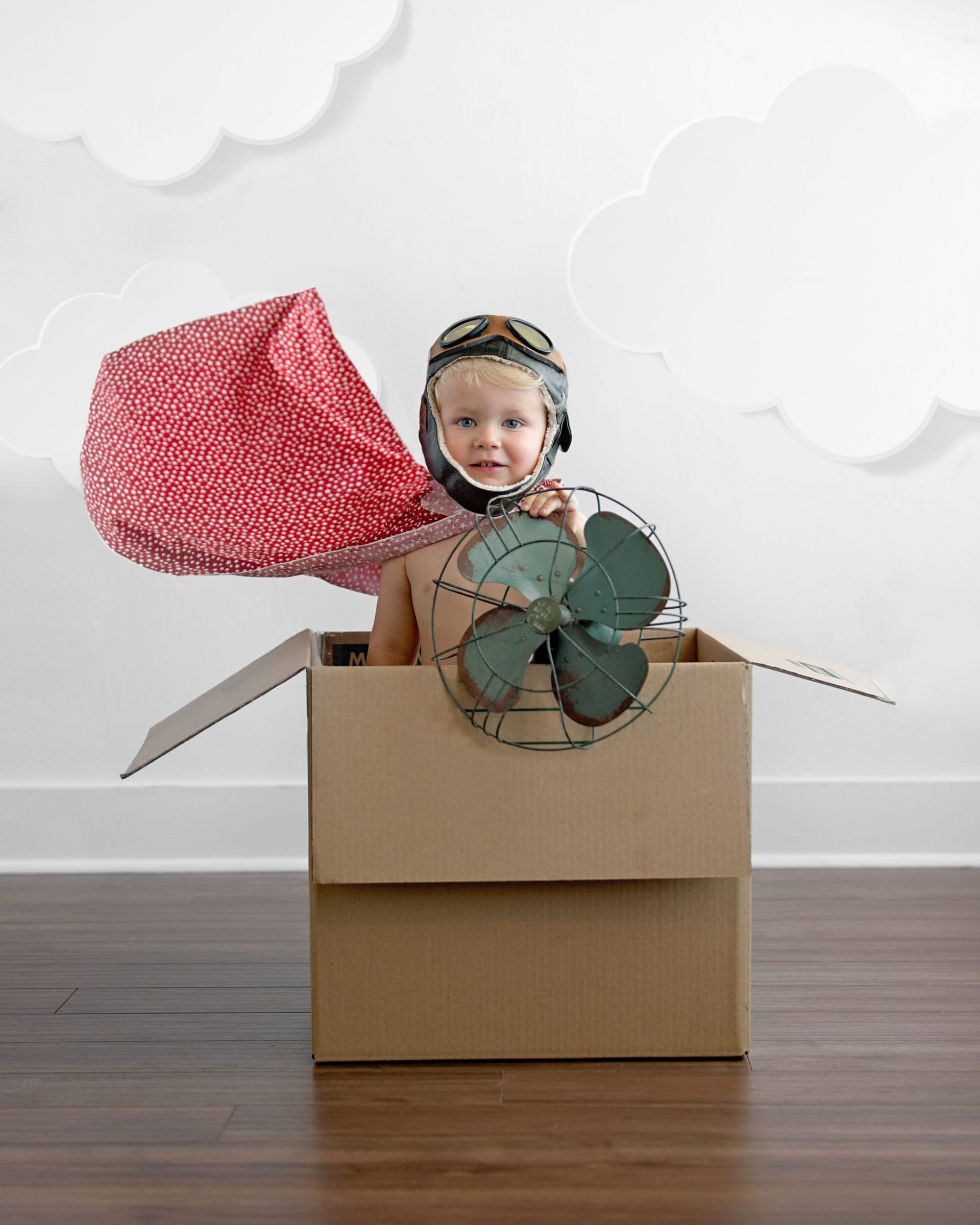 Child in cardboard box with pilot helmet and fan, red wing, smiles in front of clouds.