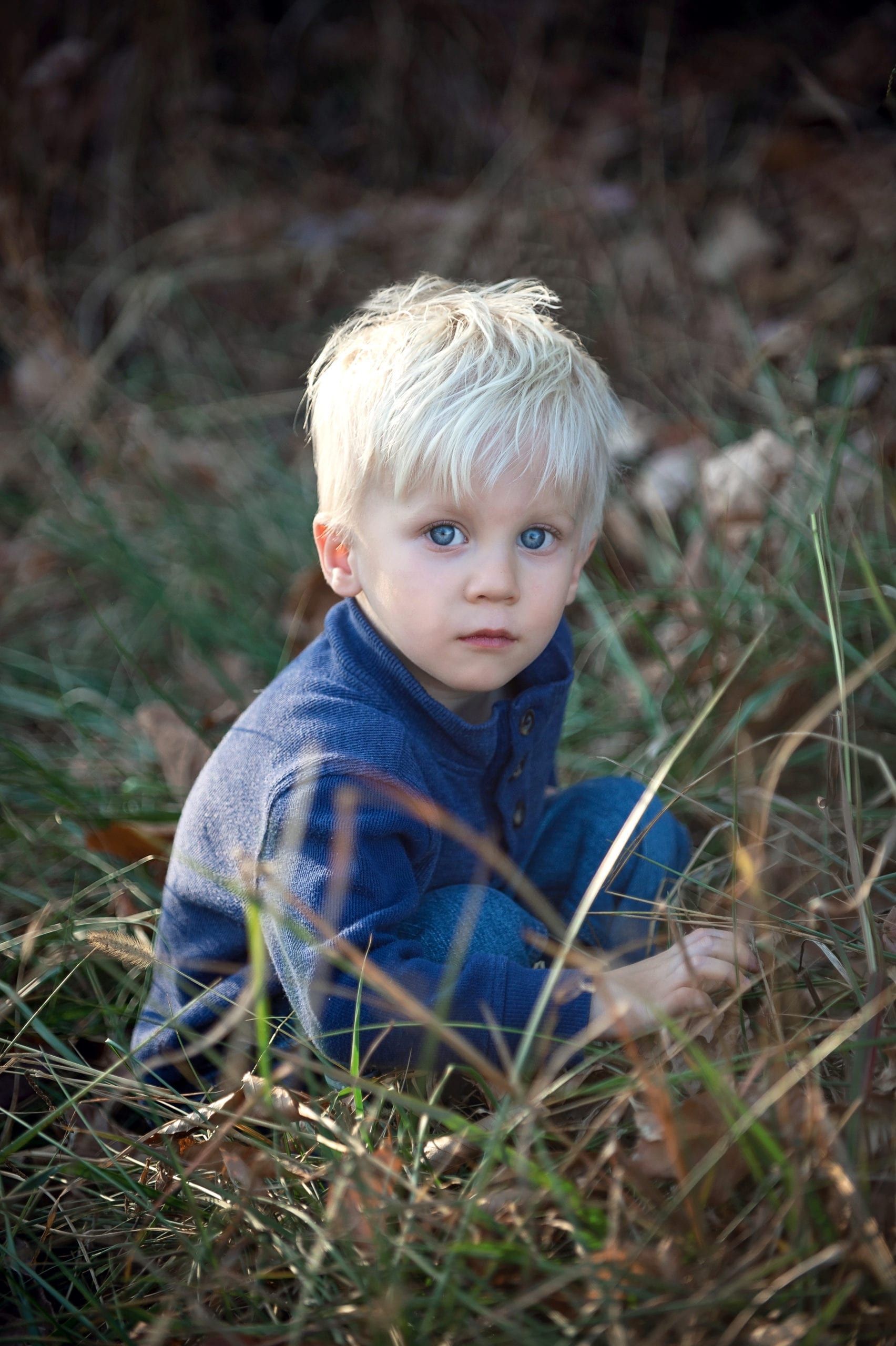Blond-haired child with blue eyes, wearing blue jacket, squatting in grass.