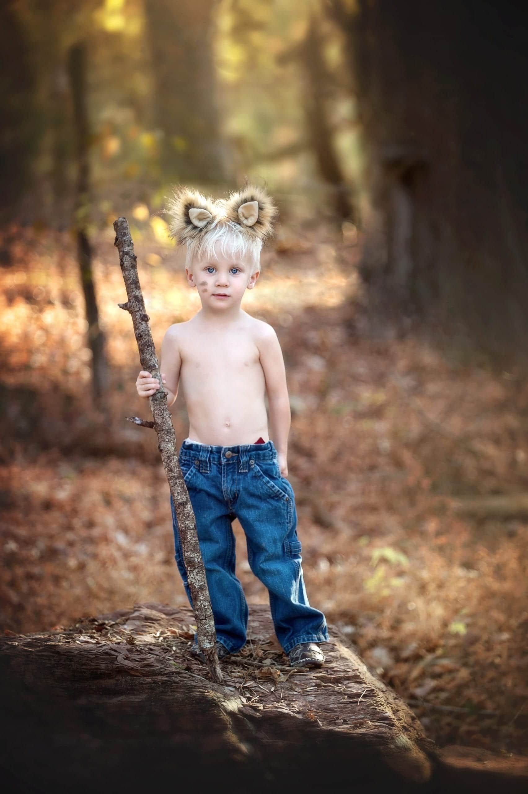 Blond child with lion ears, shirtless in jeans, holding a stick, stands on a log in the woods.