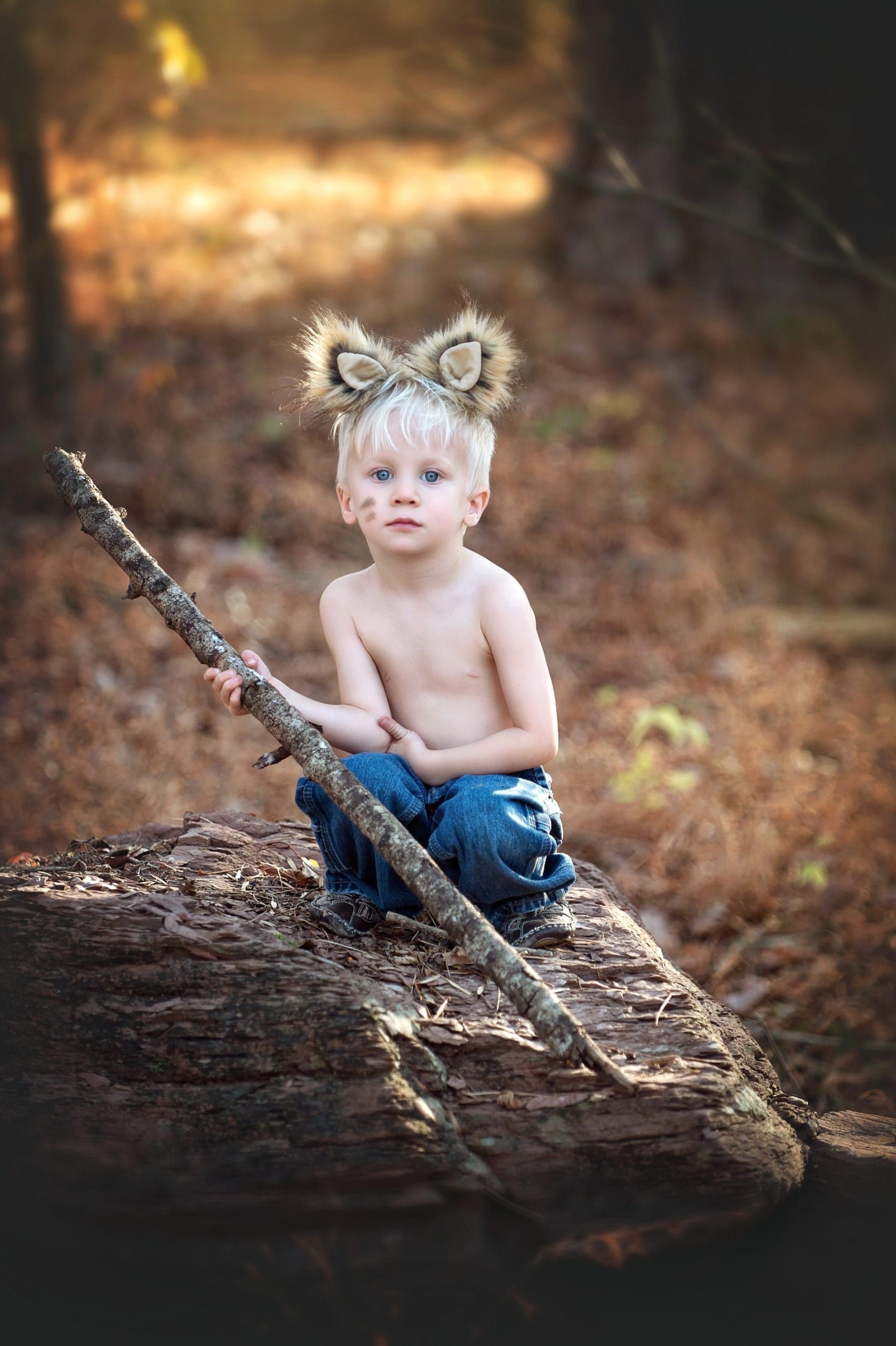 A young boy with wolf ears, shirtless, sits on a log in the forest holding a stick.