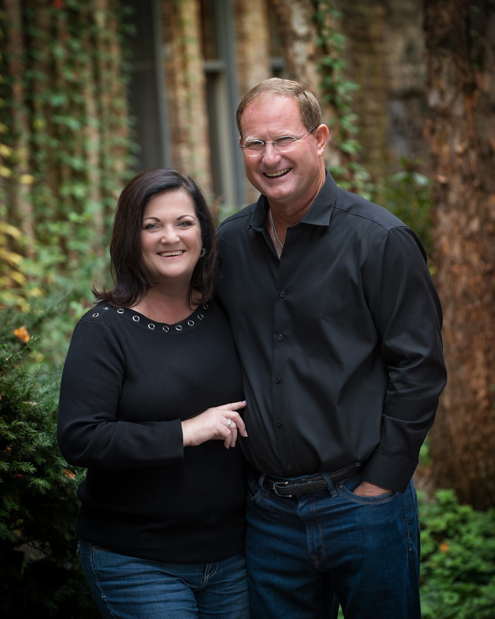 Smiling couple in front of a brick wall covered in ivy; woman in black sweater, man in black shirt and jeans.