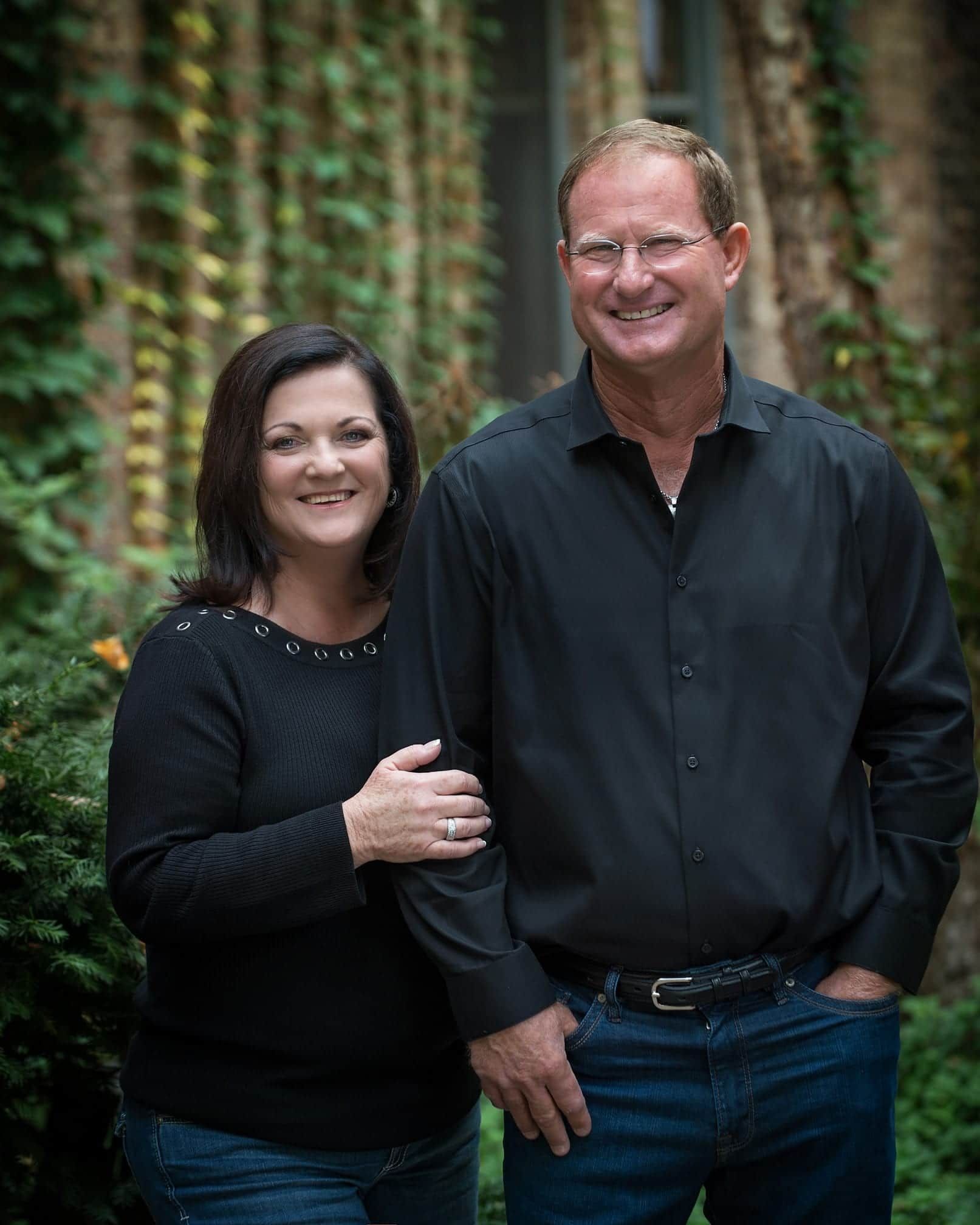 Woman in black top with arm on man's shoulder. Man in black shirt, blue jeans, smiling. Outdoors, greenery background.
